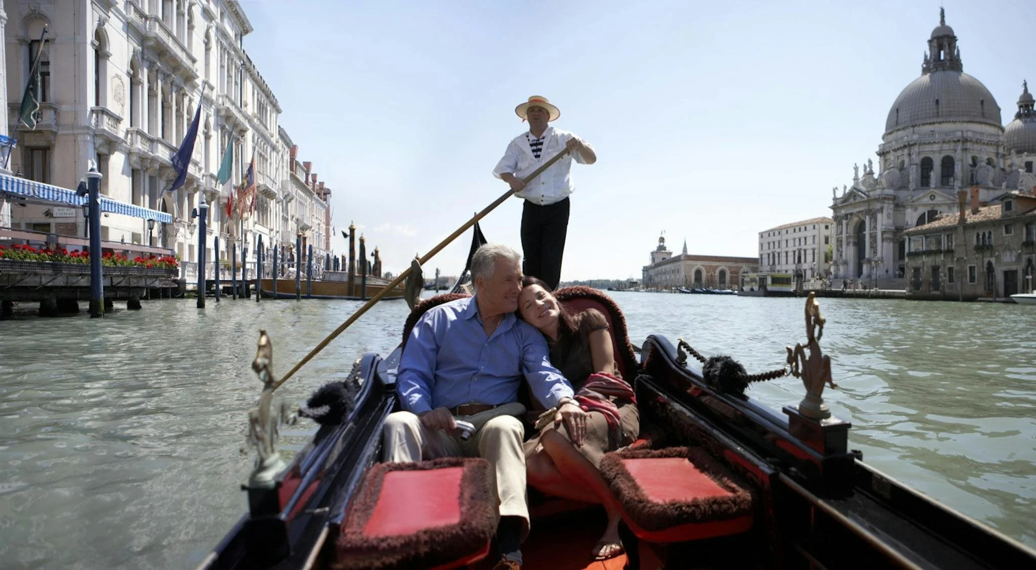 A couple sits enjoyably together on a gondola ride in Venice, Italy, with the grand basilica visible in the background. The gondola is steered by a gondolier in traditional outfit.