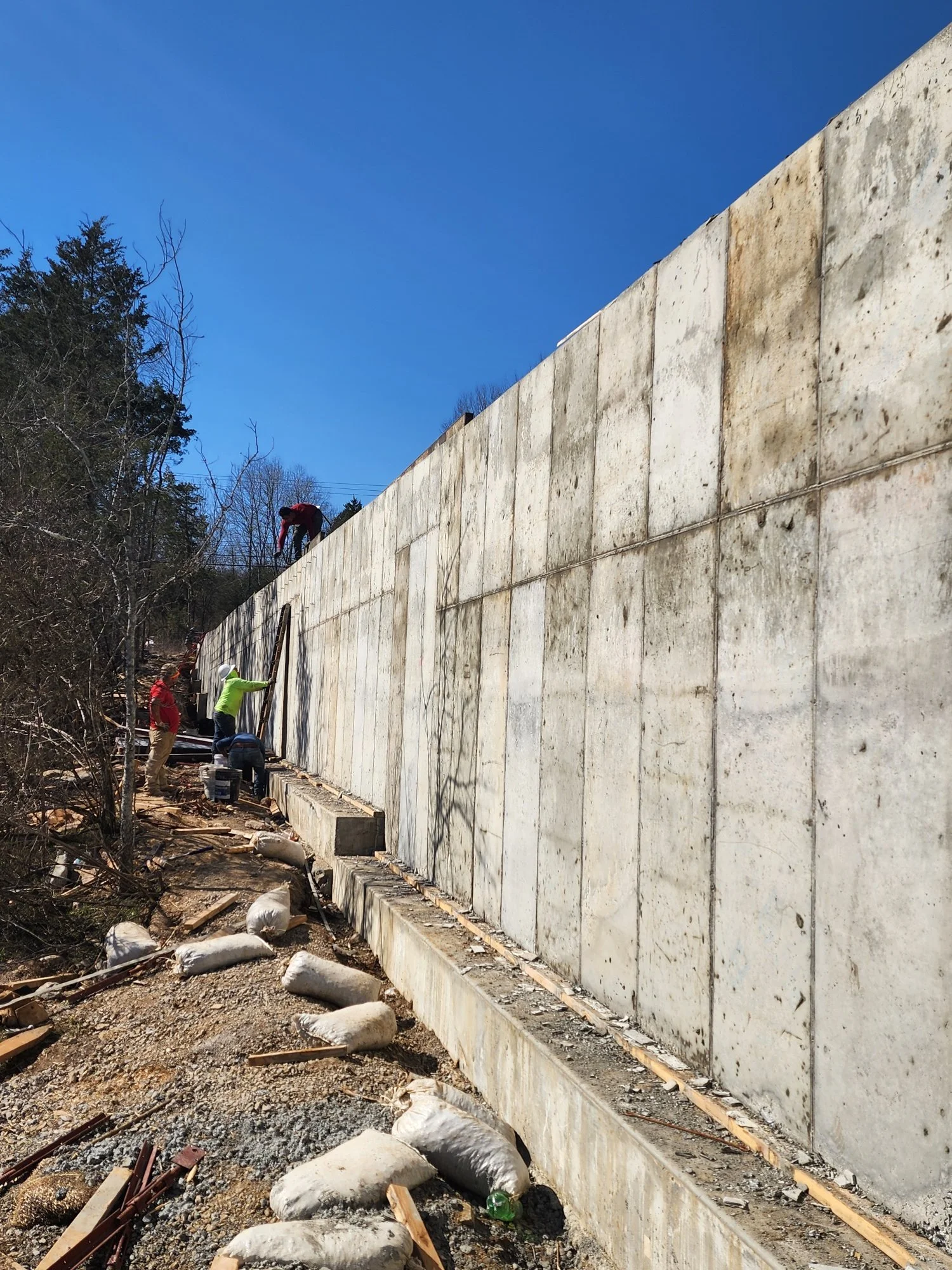 Construction workers building a concrete wall outdoors on a sunny day, with trees on the left and a clear blue sky above.