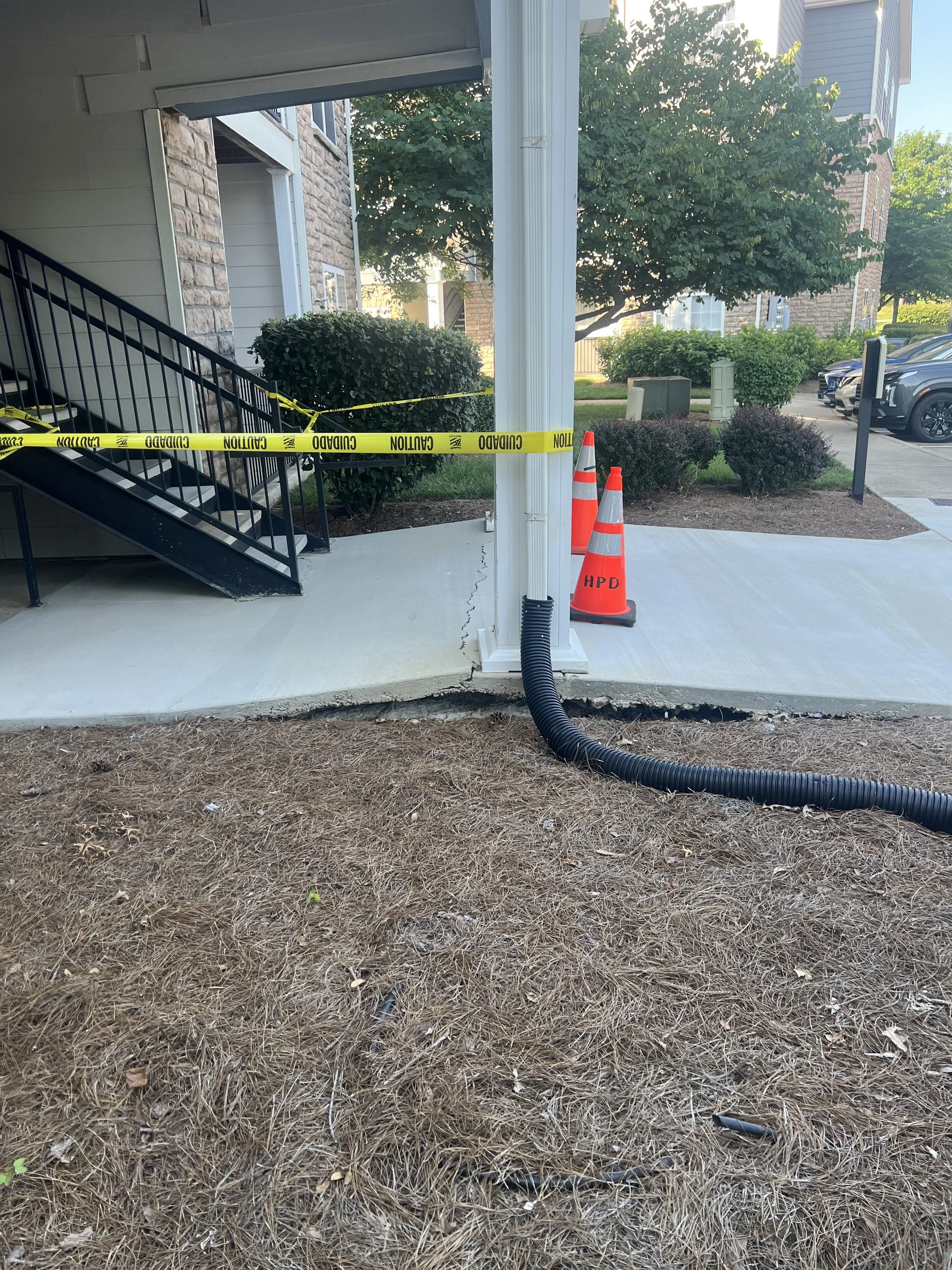 Construction area with caution tape, orange cones, and a large black drainage pipe along the ground outside a building with shrubbery and parked cars in the background.