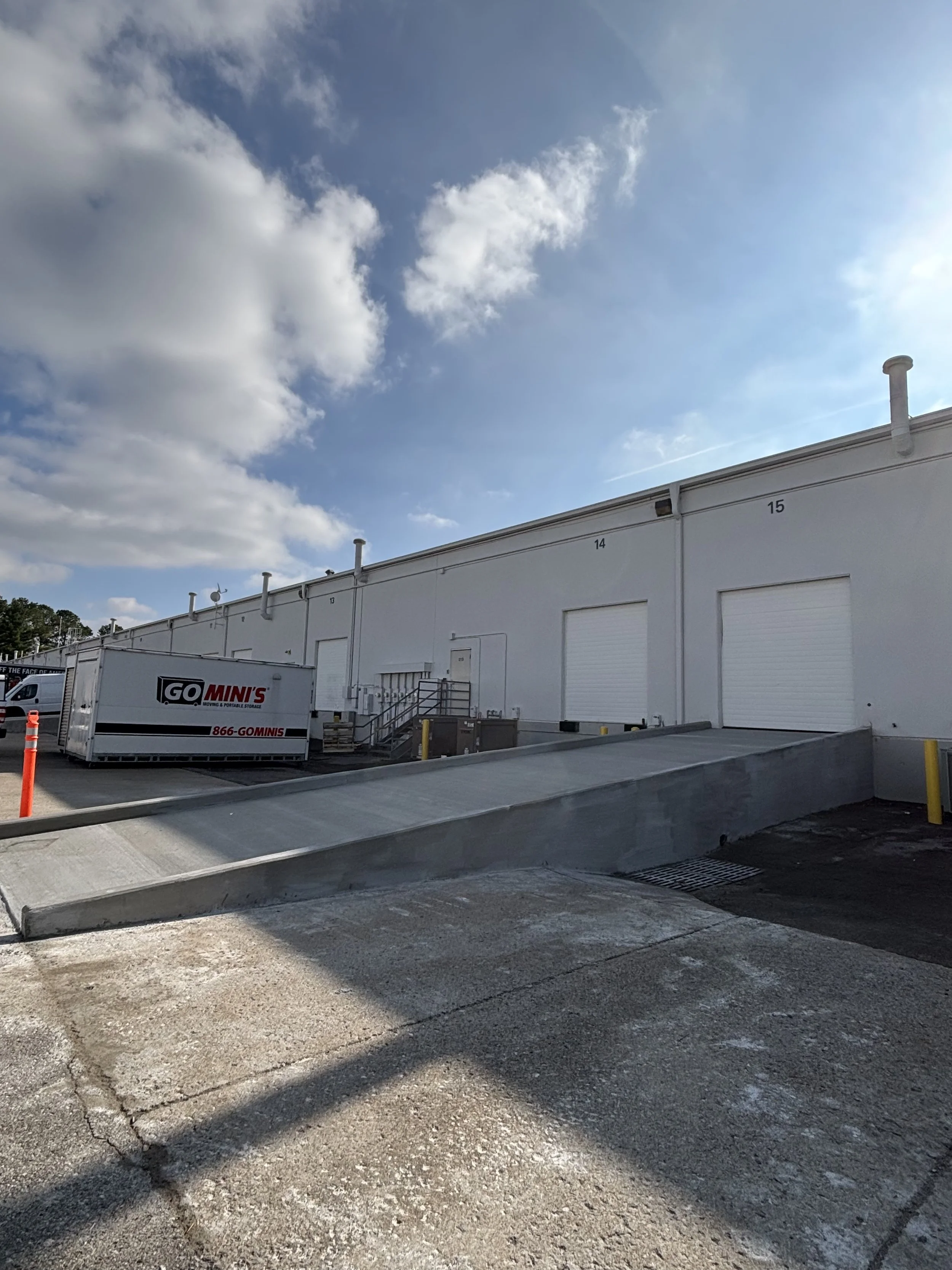Loading dock with a sloped concrete ramp in front of a white warehouse building, two large roll-up doors, and a GO MINI'S moving container nearby, under a partly cloudy sky.