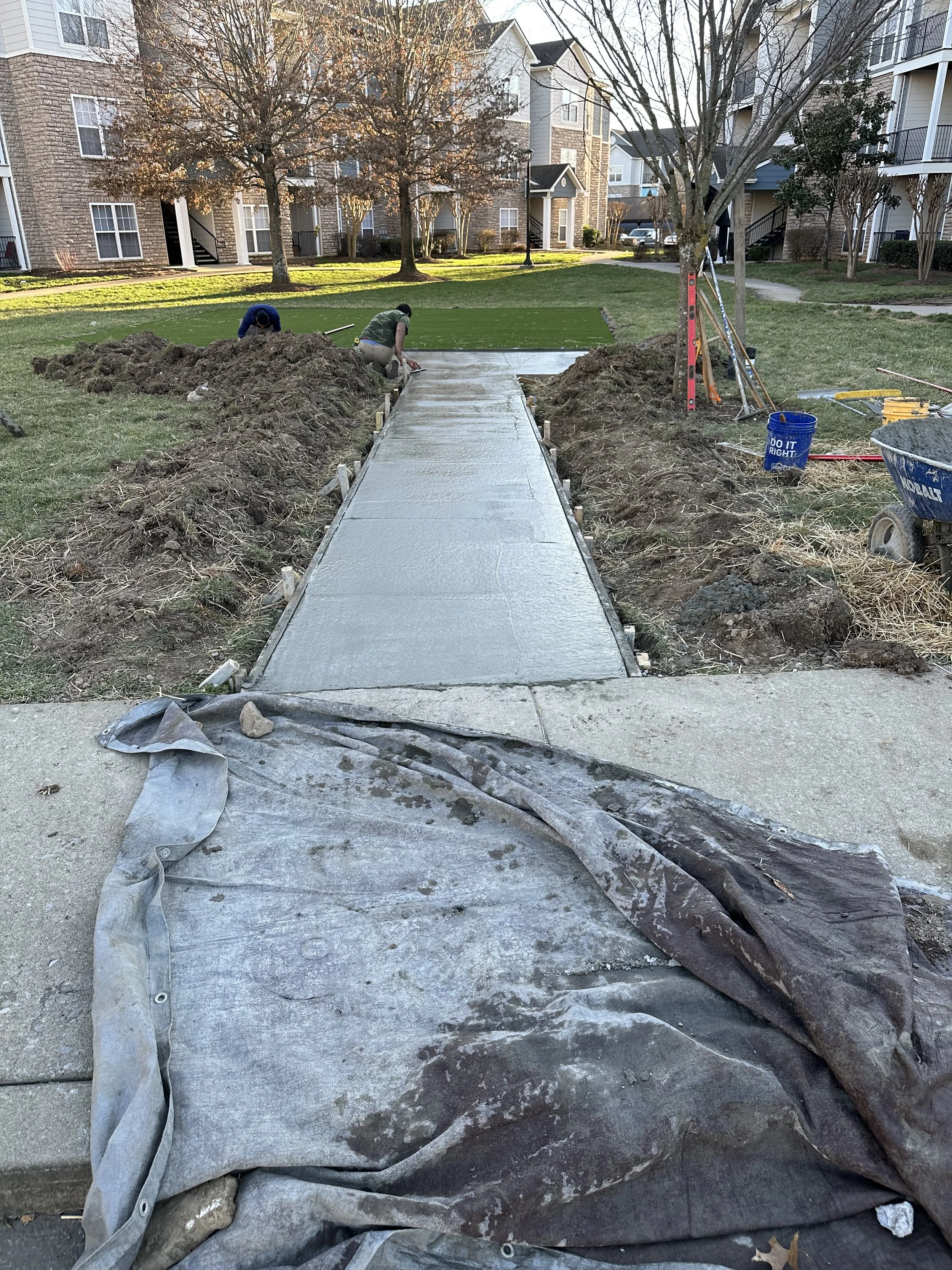 Construction workers pouring concrete to finish a new sidewalk in a residential area with apartment buildings and trees in the background.