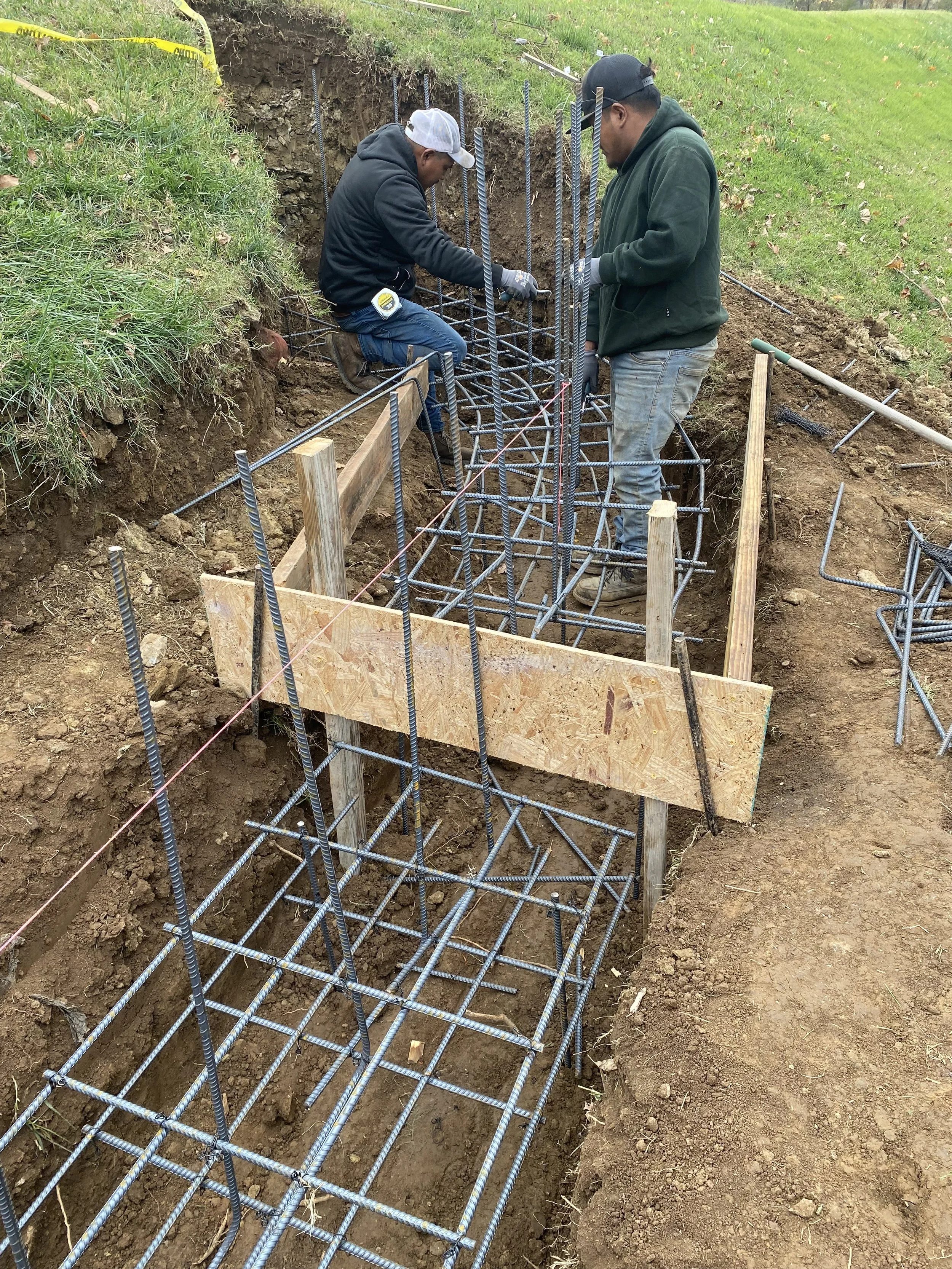 Two workers installing rebar reinforcement in a concrete foundation trench outdoors, with wooden formwork and yellow caution tape nearby.