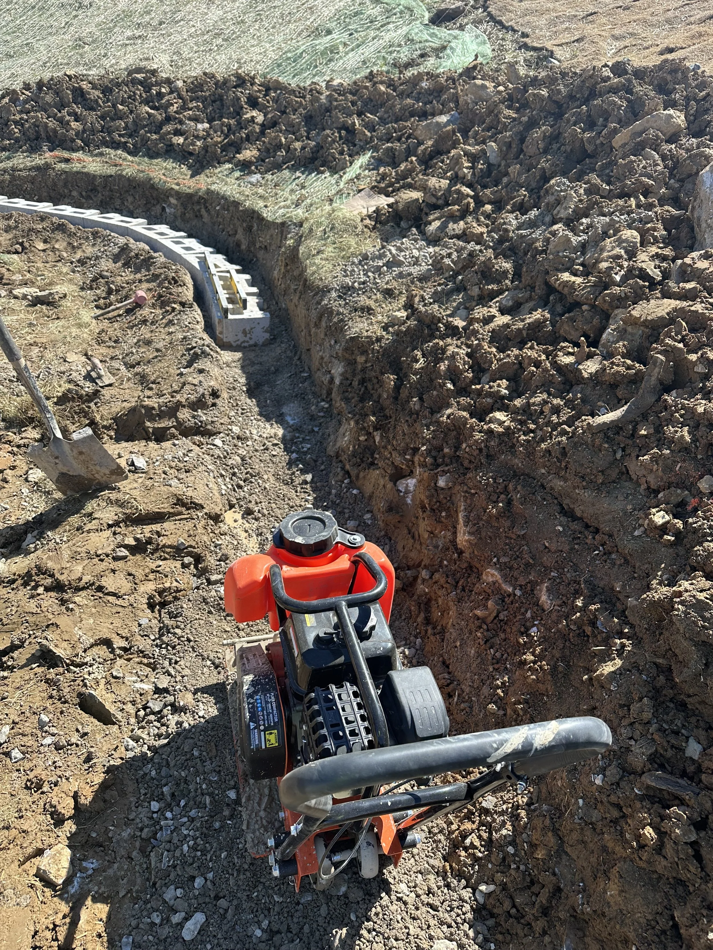 View of a construction site showing a small dirt trench with a curved concrete block wall in progress and a red plate compactor machine on the ground. There is loose dirt, some tools, and a shovel nearby.