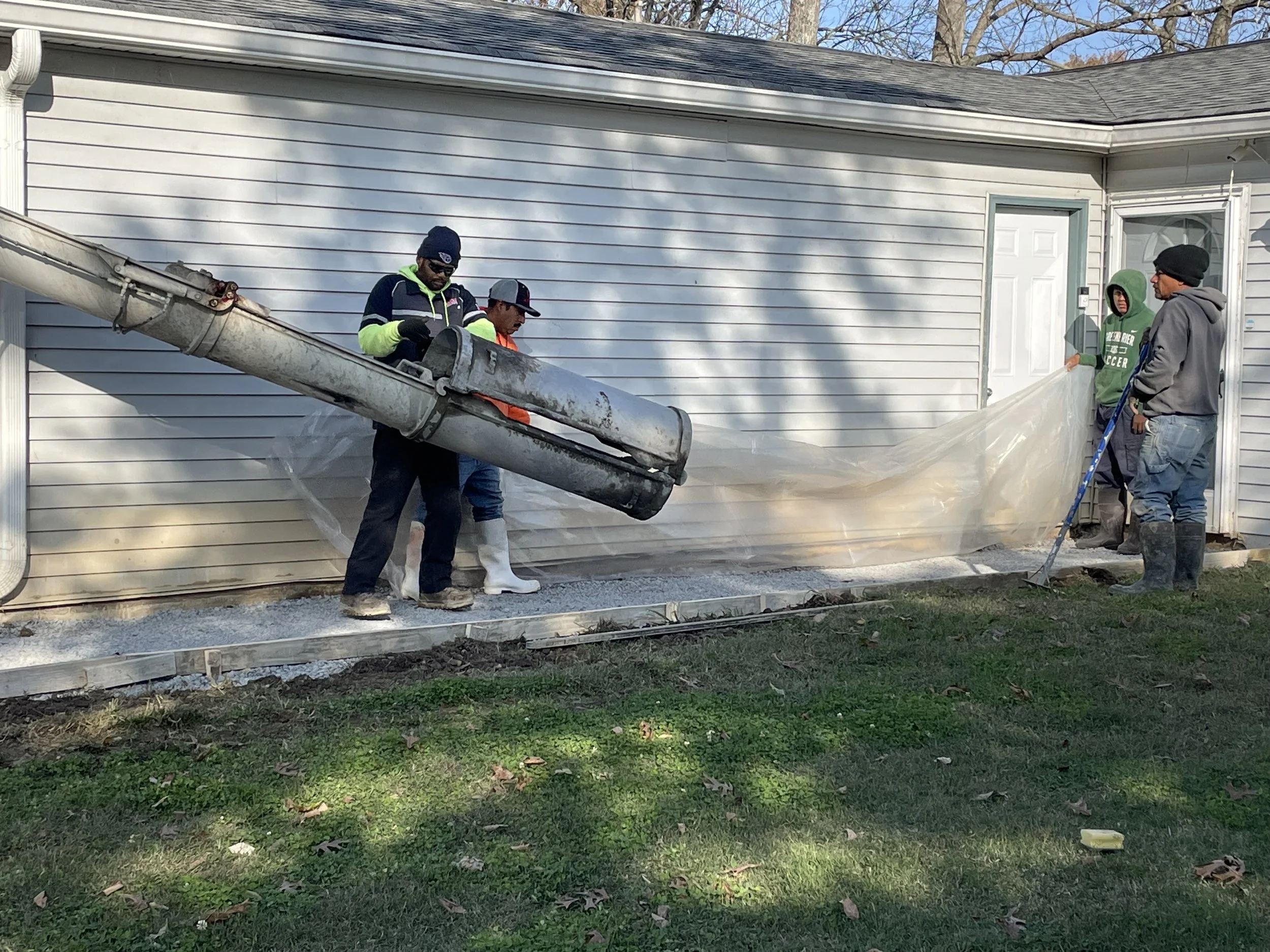 Group of five workers installing foundation or sidewalk, with gravel and plastic sheeting in front of house, using a concrete pump.