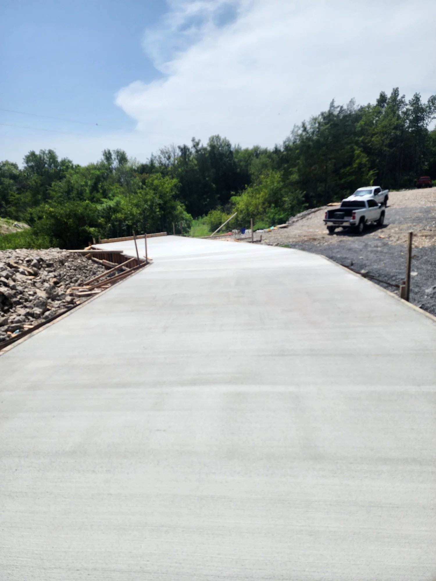 Newly poured concrete road with construction barriers on the sides and parked trucks in the background surrounded by green trees and an overcast sky.