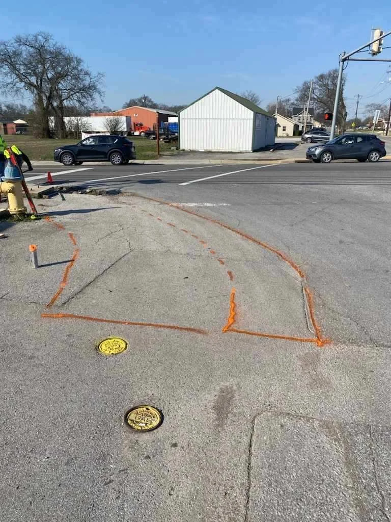 Roadwork with orange safety markings on sidewalk near a cross street, with water valve covers and traffic lights in the background.