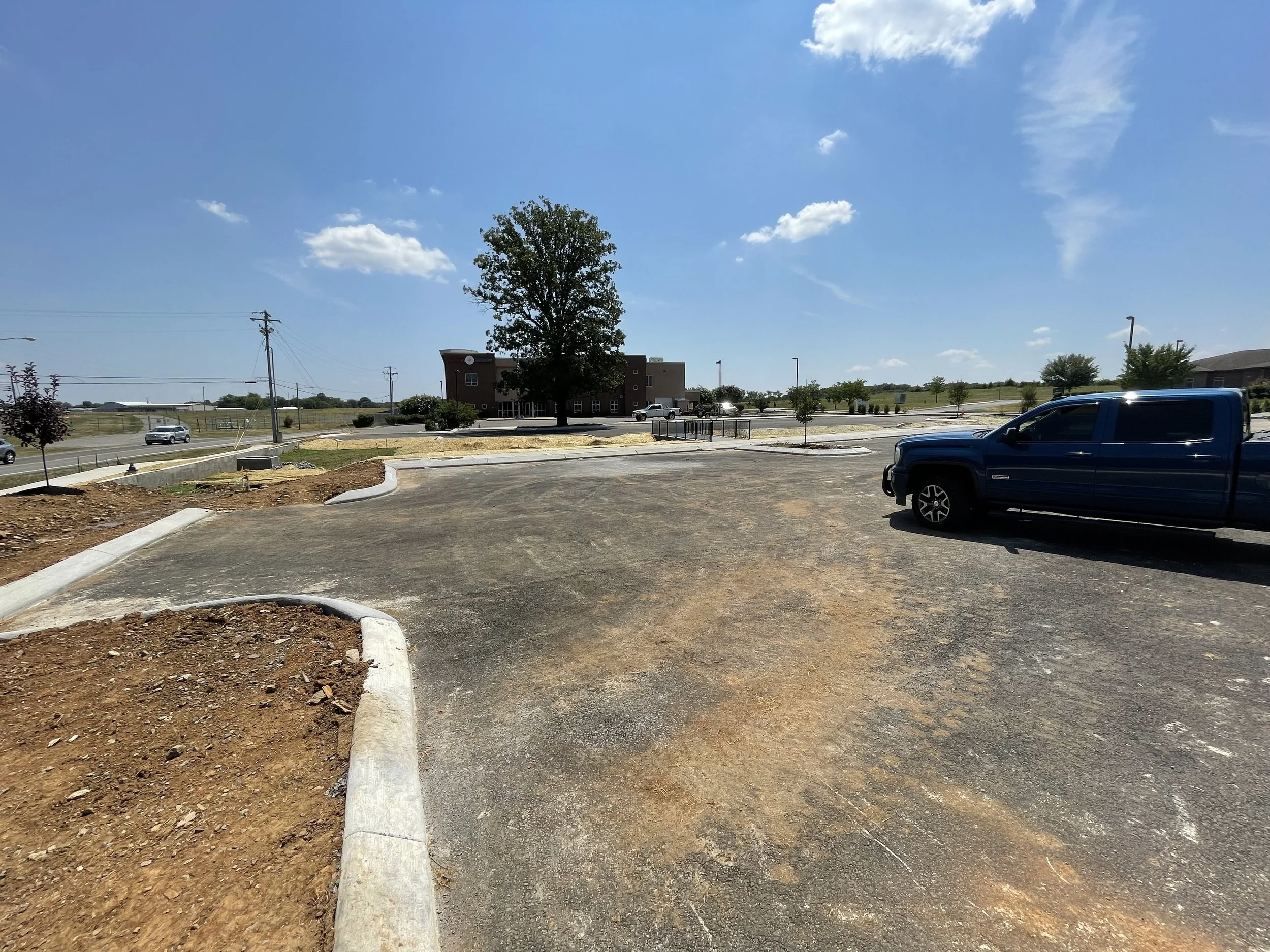 Partially paved parking lot with a black truck on the right and a building with trees in the background. Under construction or landscaping in the foreground with bare soil and concrete curbs.