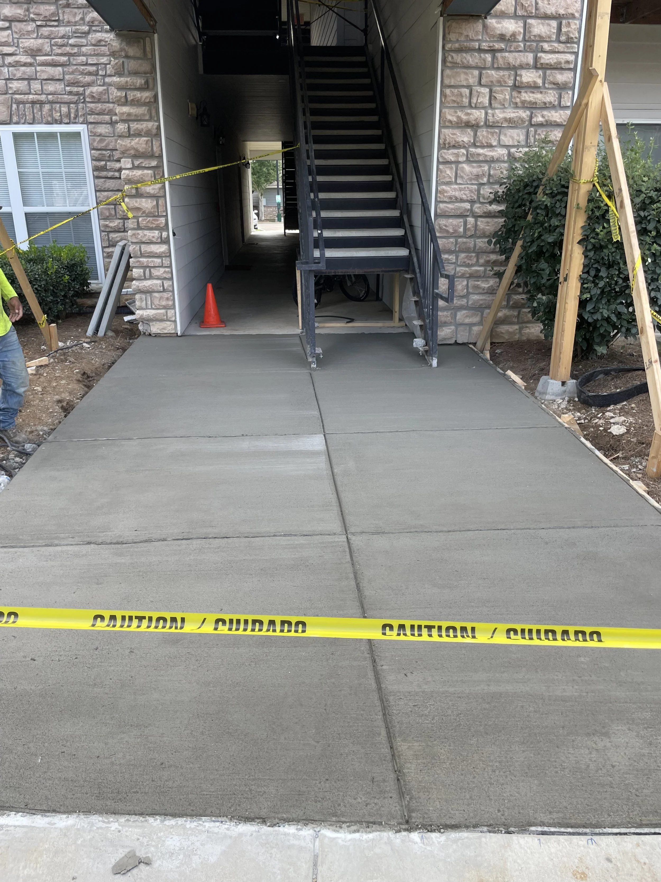 Freshly poured concrete sidewalk in front of a building with caution tape, construction cones, and ongoing landscaping work.