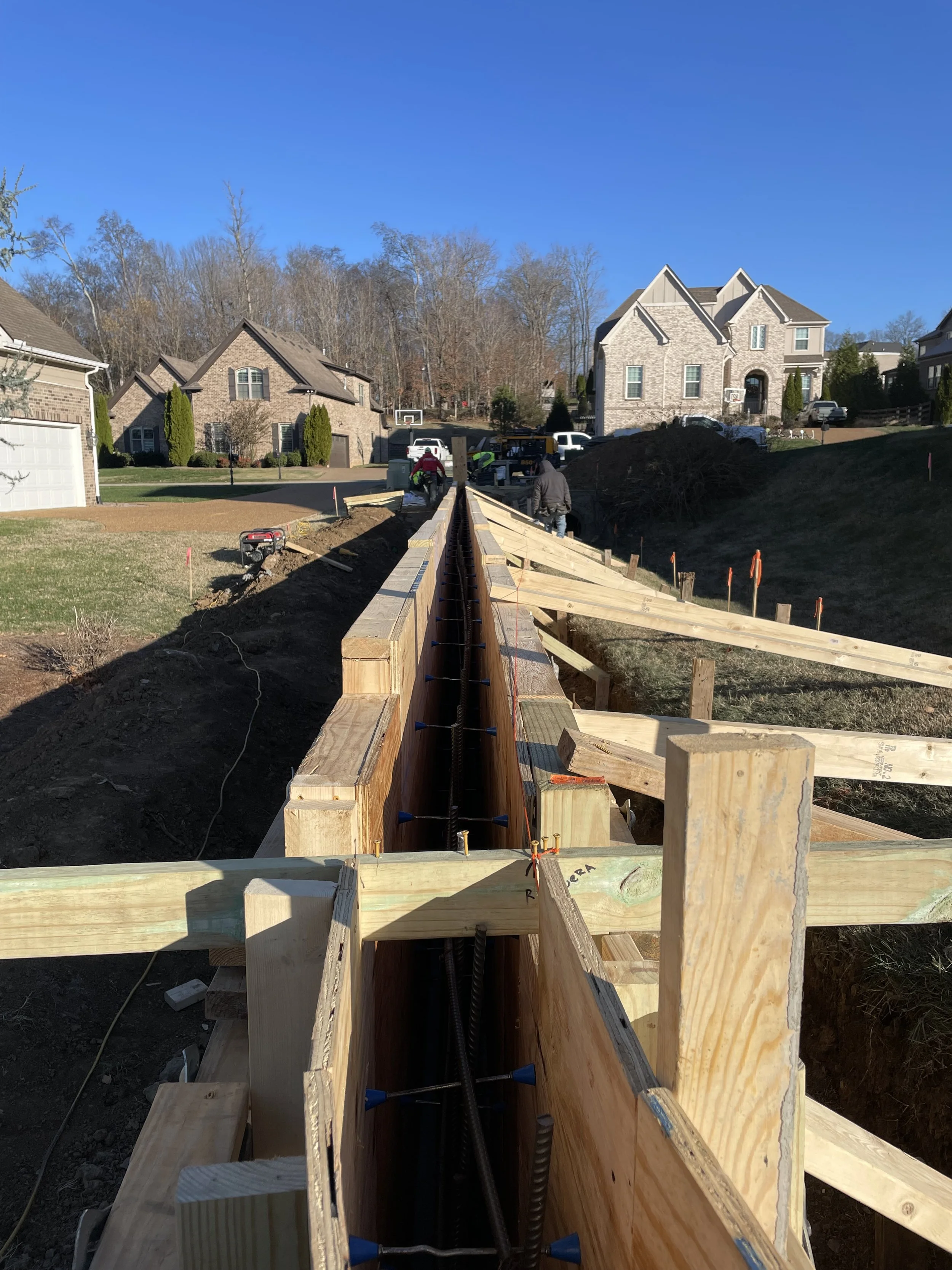 Construction site with wooden framework for a new structure in a residential neighborhood, with houses and trees in the background.