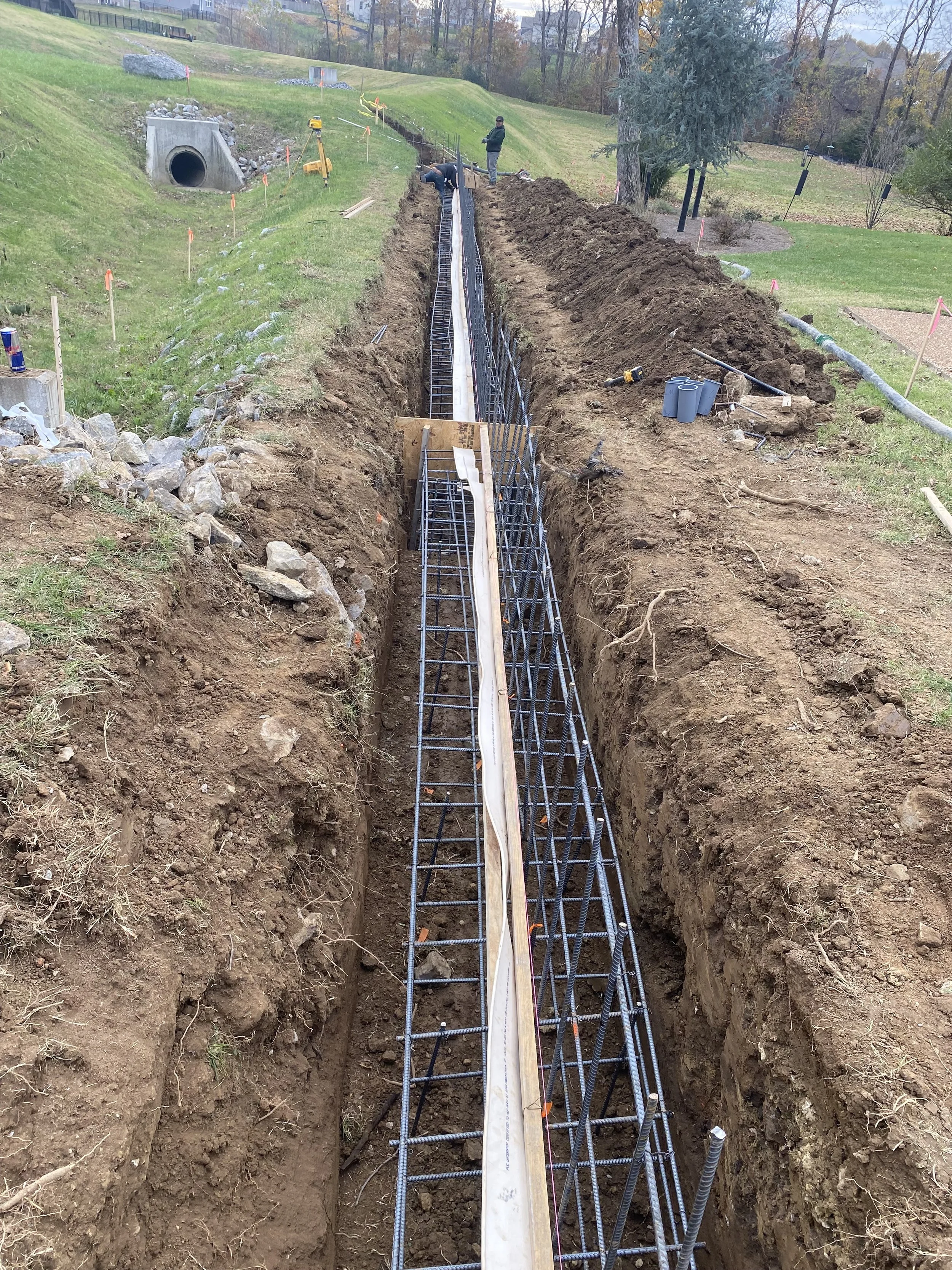 Construction workers install rebar in a long trench for a concrete foundation on a hillside with grassy areas and trees in the background.