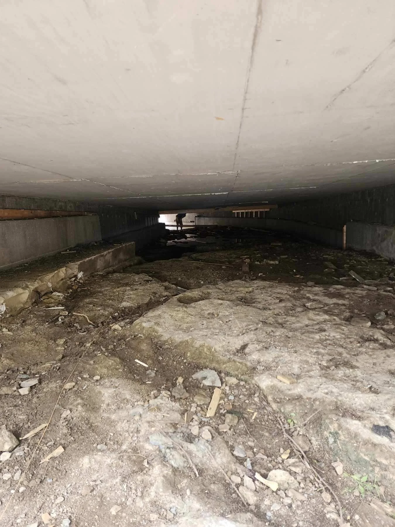 Underneath a building or bridge, with uneven dirt and rocks on the ground and a person in the background working or walking.