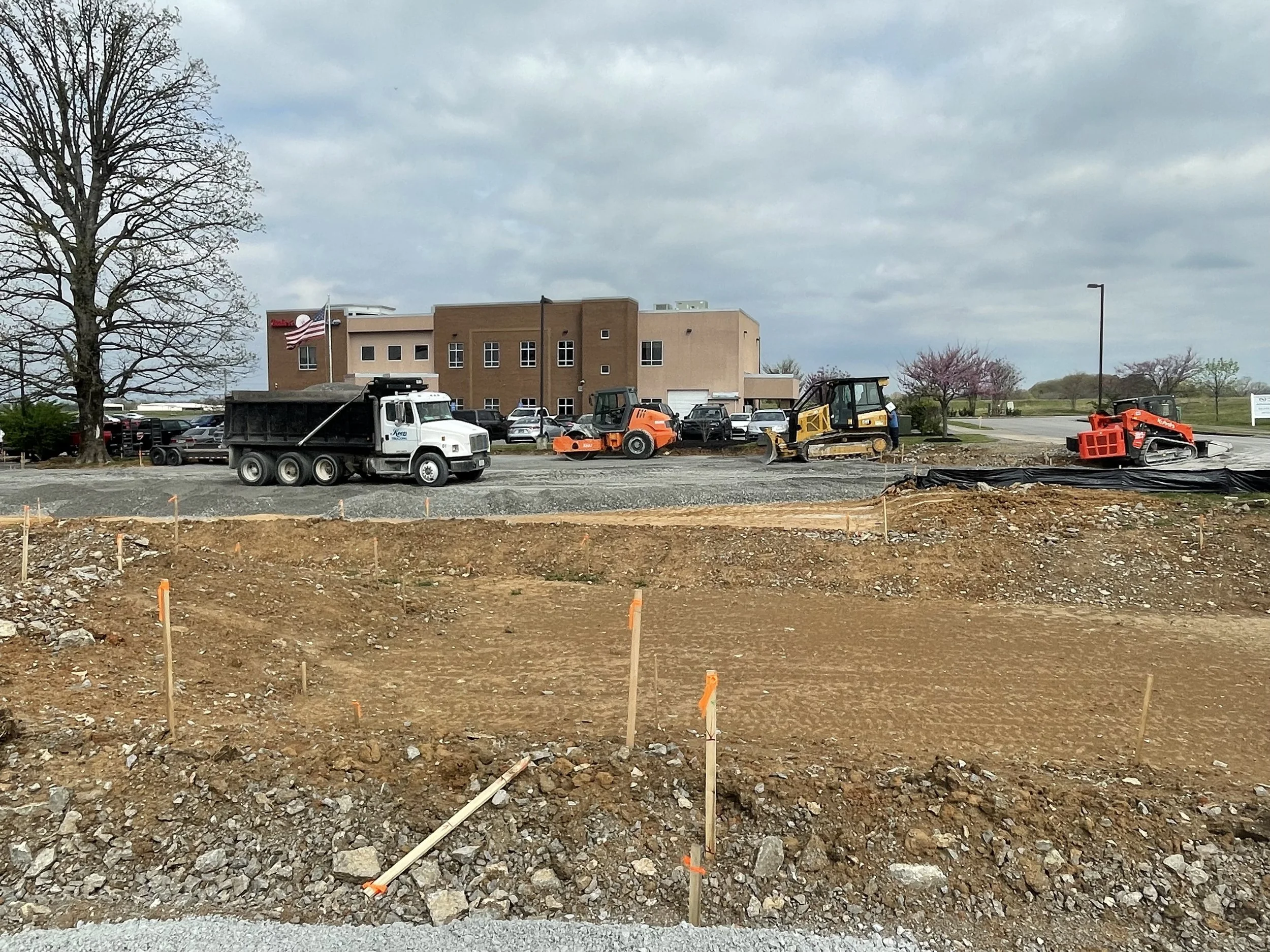 Construction site with dirt and gravel, construction equipment including a roller and paver, parking lot with cars, and a building with an American flag in the background.