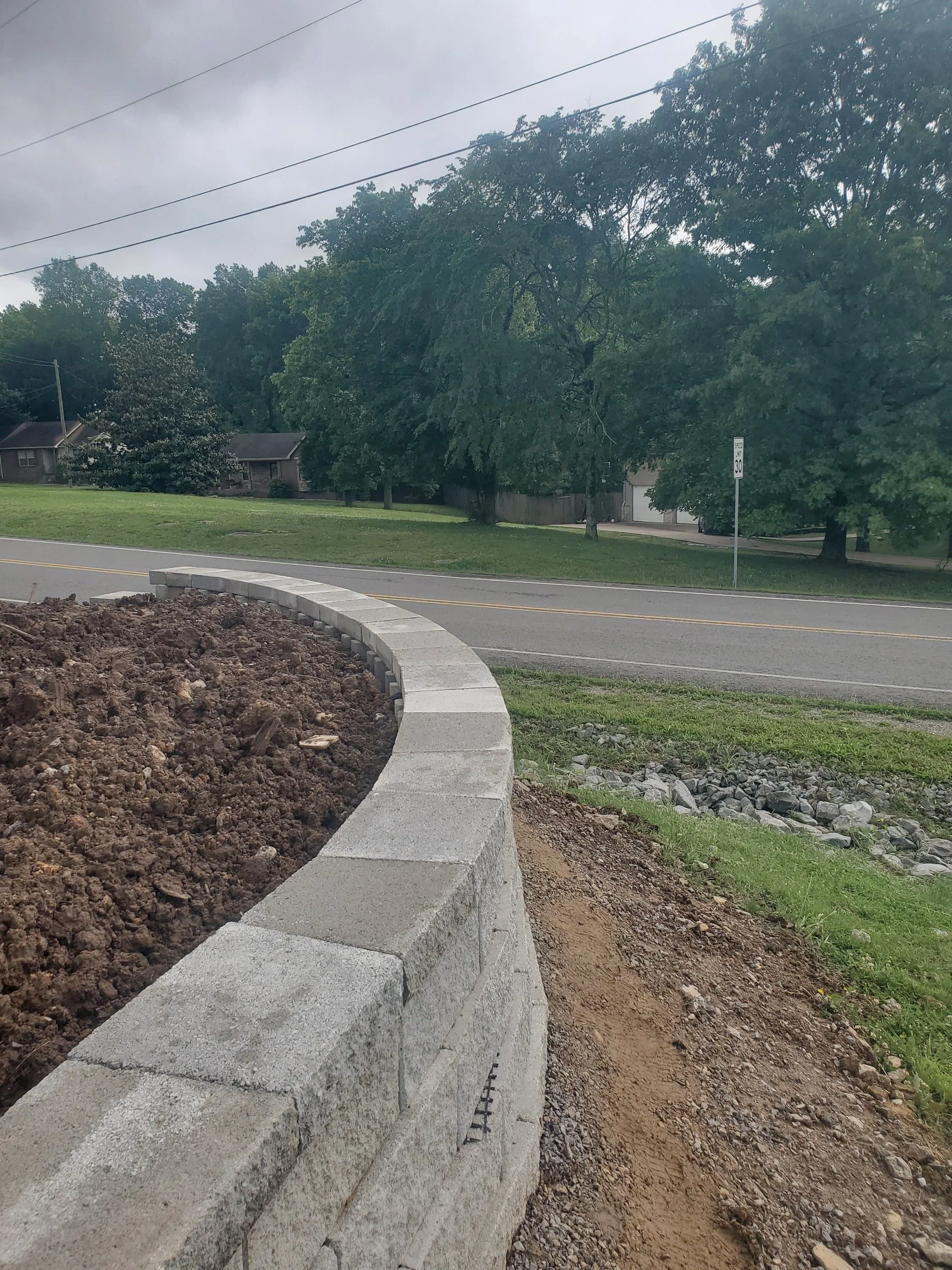A curved stone brick retaining wall with soil behind it, next to a grassy area and a two-lane road. There are trees, houses, a speed limit sign, and overhead power lines in the background.
