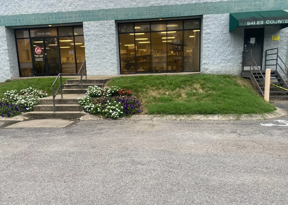 Exterior of a building with glass windows, small steps leading to the entrance, flower beds with white, purple, and red flowers, grassy area, and a sign indicating 'Sales Counter'