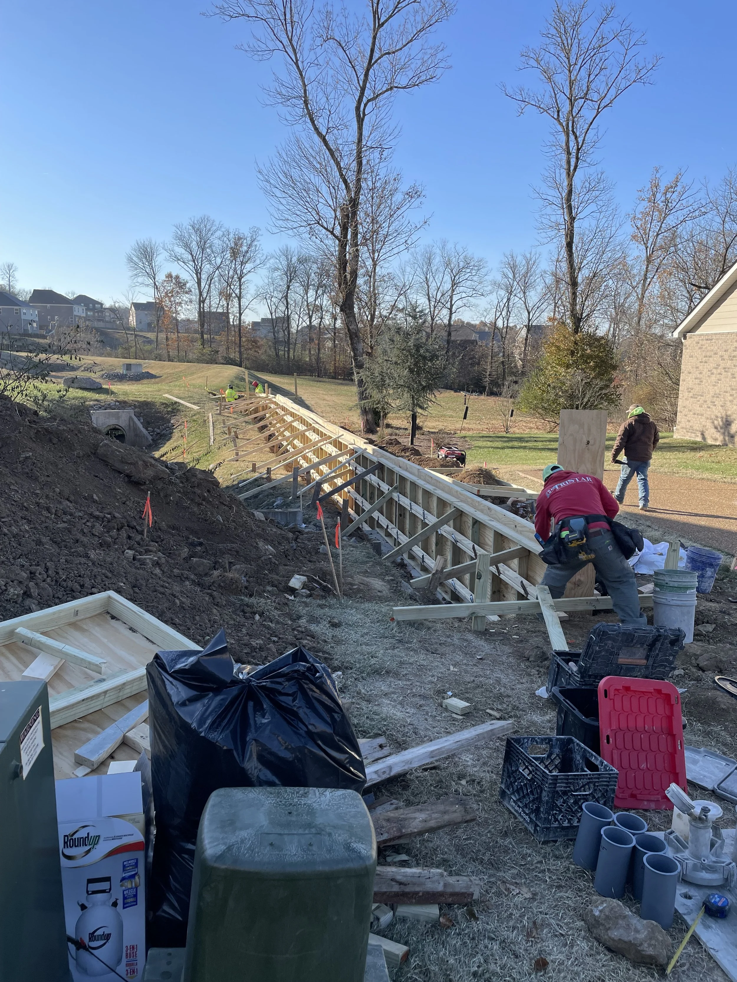 Construction workers building a wooden retaining wall on a sloped backyard, with tools, materials, and equipment scattered around.