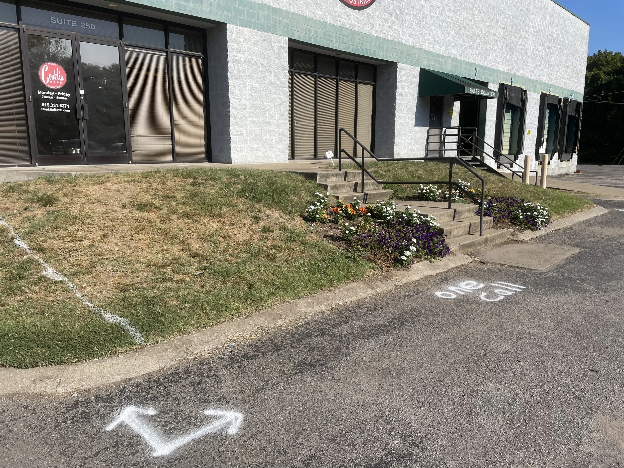 Parking lot with arrow and 'NO SIGN' painted on the ground, front of a commercial building with stairs and flower bed.