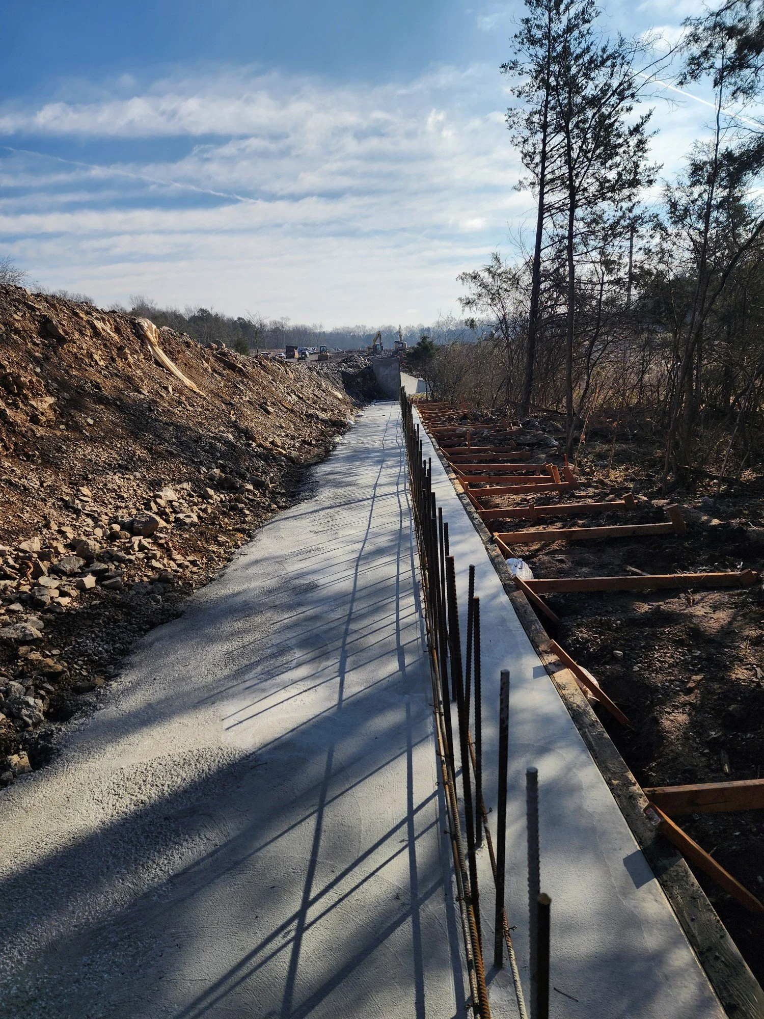Newly poured concrete sidewalk along a construction site with steel rebar and wooden formwork, surrounded by dirt and trees, under a partly cloudy sky.