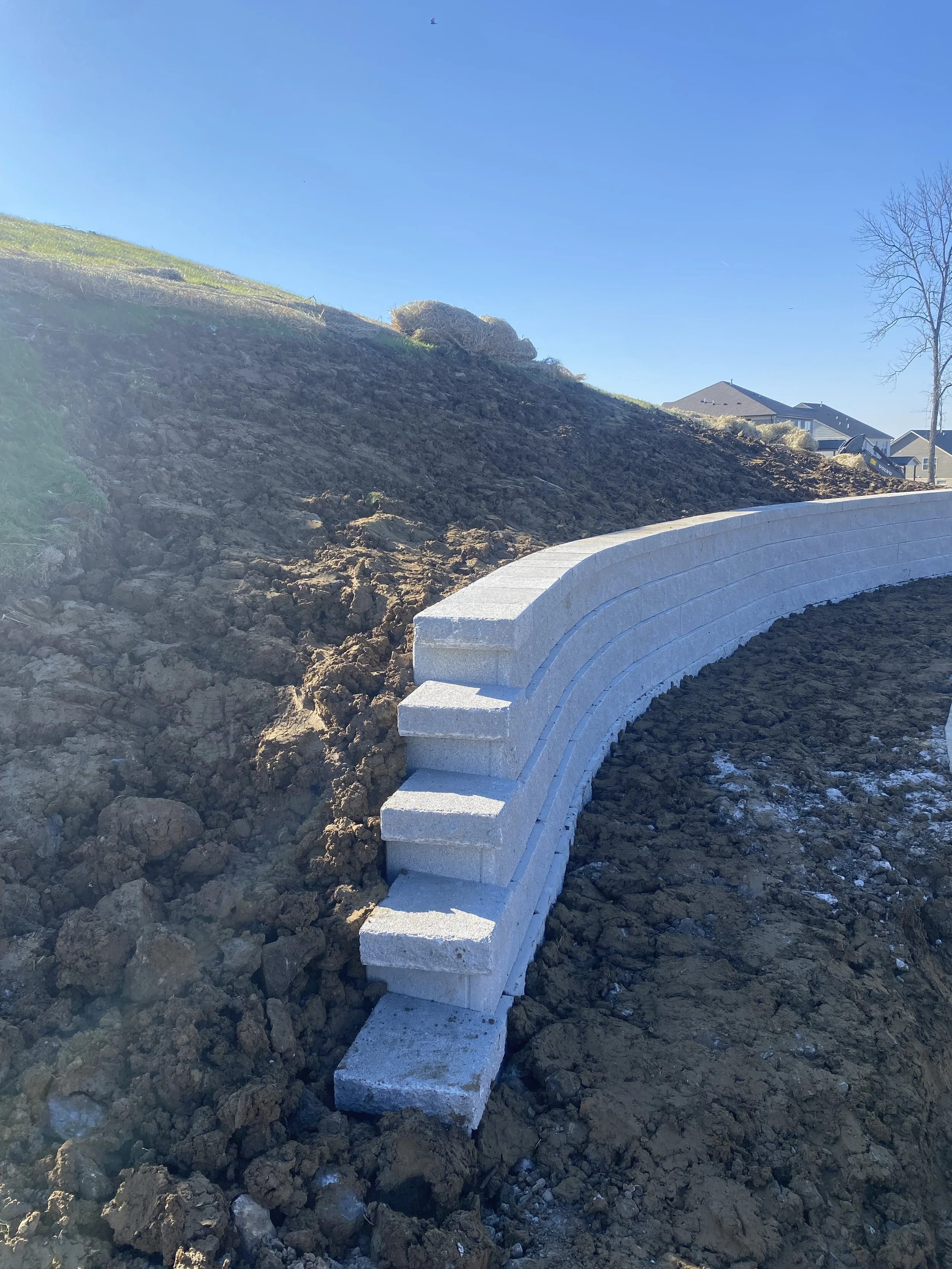 A curved retaining wall made of concrete blocks on a hillside with bare soil and a few houses visible in the background.