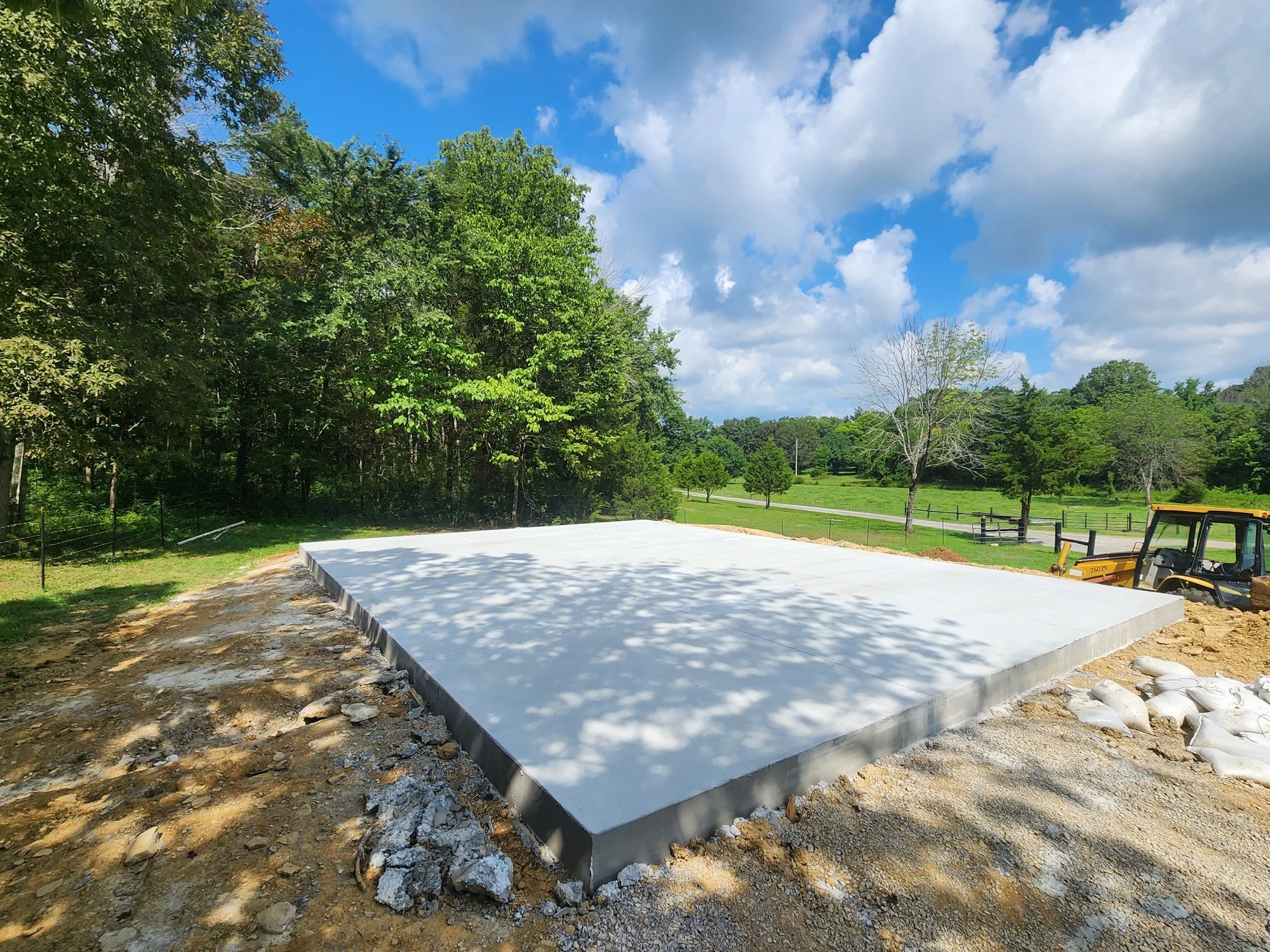 Freshly poured concrete slab in a rural outdoor setting with trees and green fields in the background, a construction vehicle nearby.