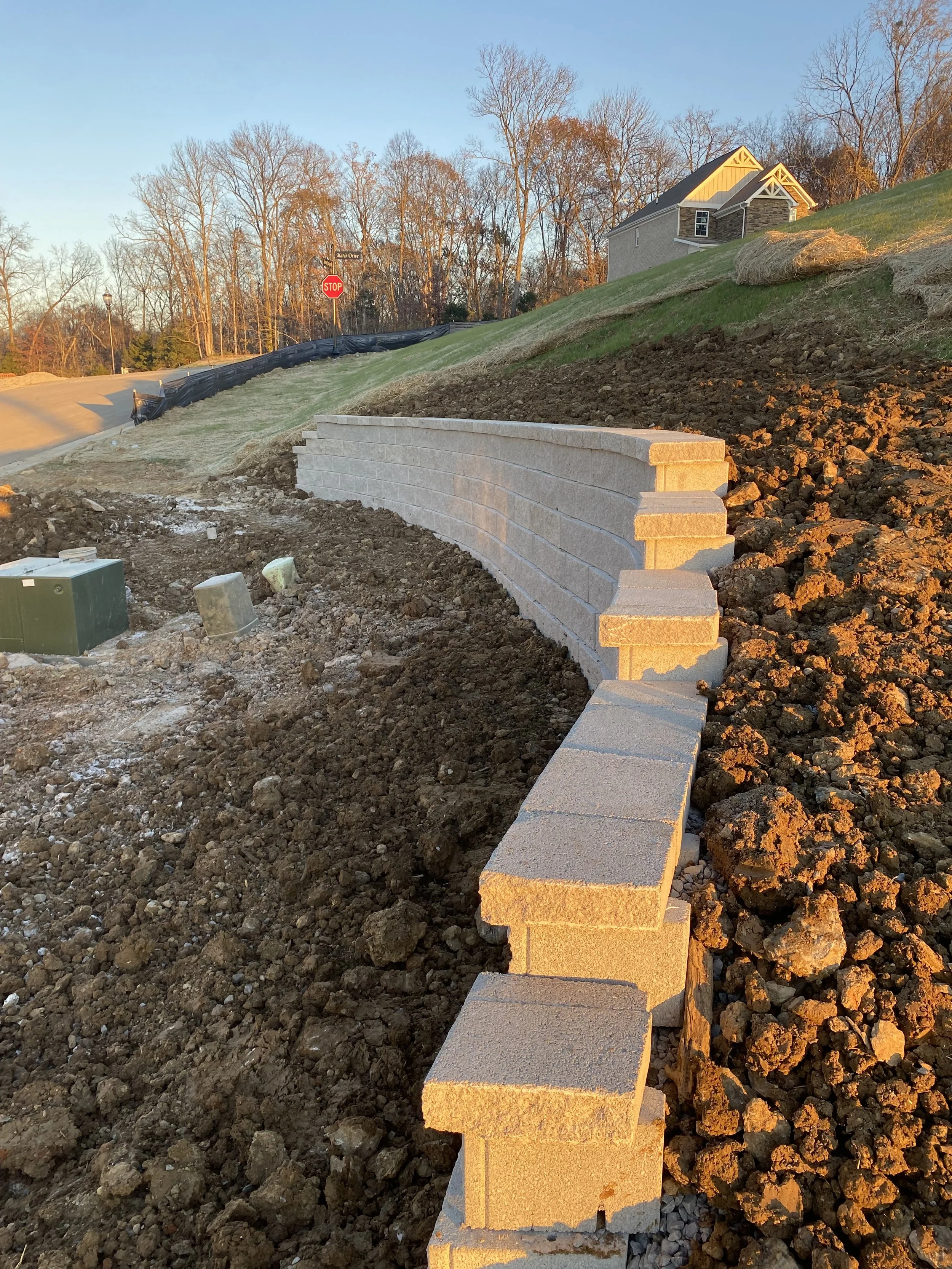 Construction site with a new concrete block retaining wall curving along a hillside in sunlight, with soil disturbed around it and houses in the background.