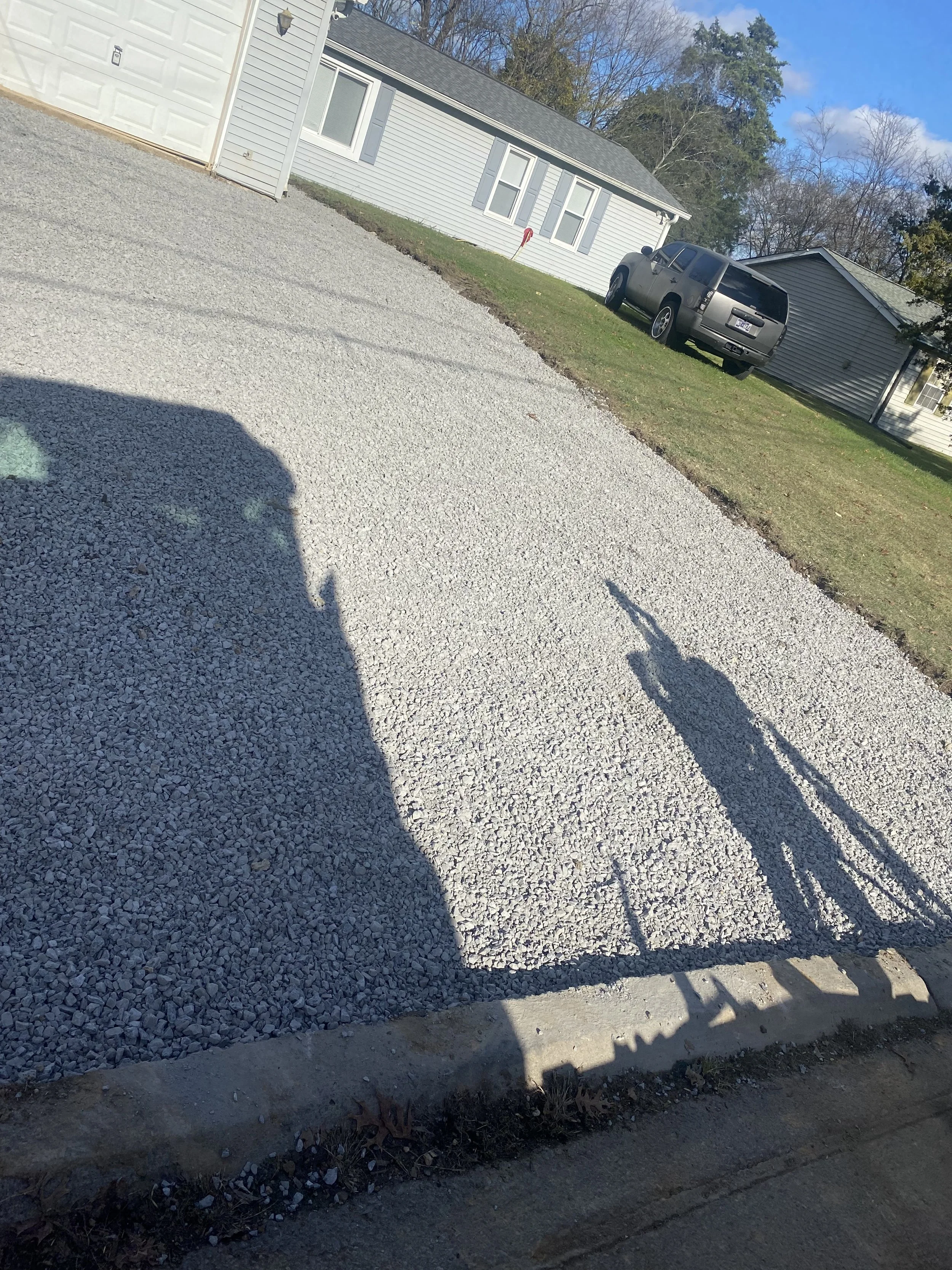 A gravel driveway with a shadow of a person taking the photo. In the background, there are two houses, one with gray siding and a black SUV parked in front of it. The sky is blue with a few clouds.