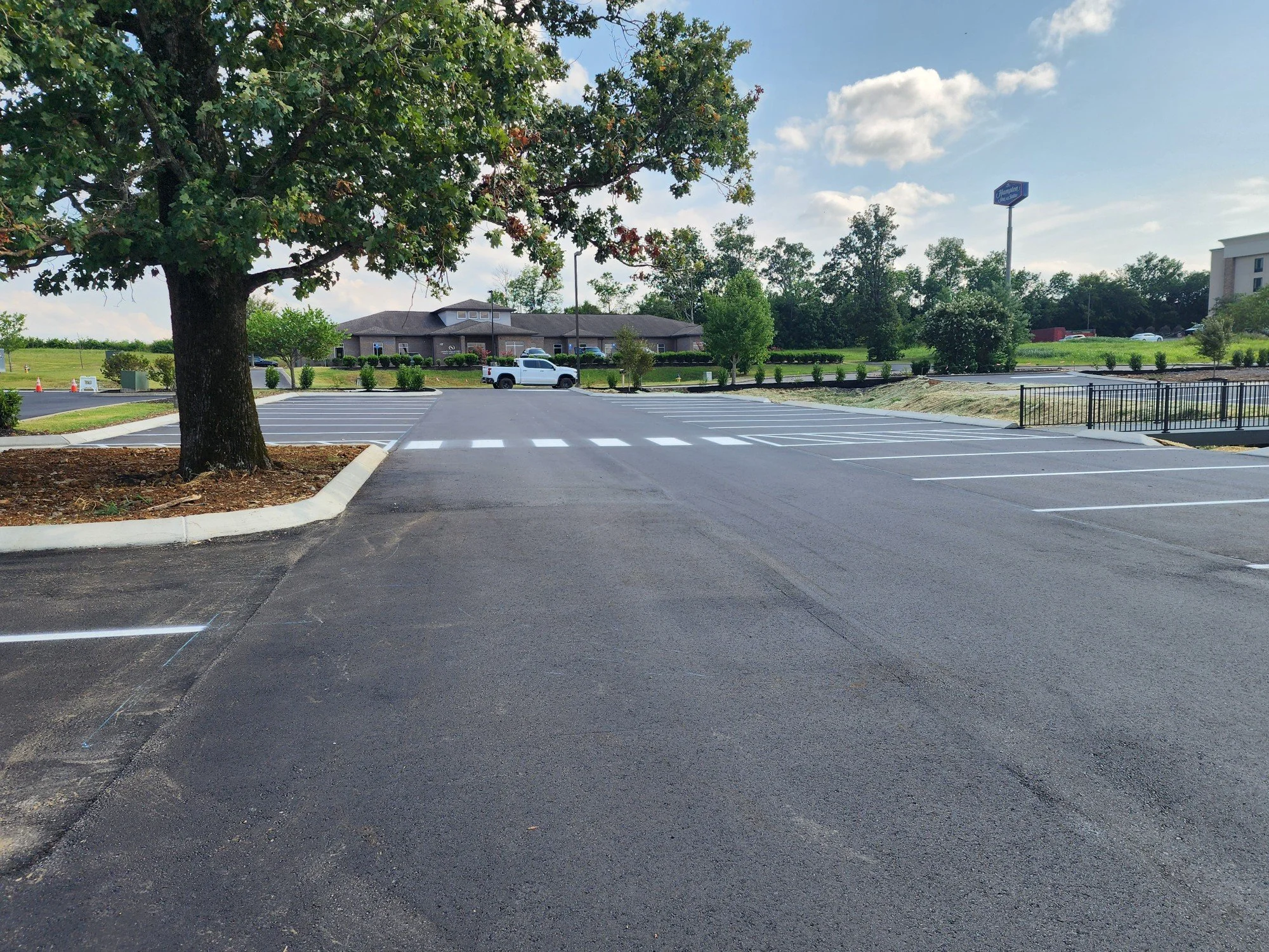 Empty parking lot with freshly painted white parking lines, a large tree on the left, a white truck in the distance, and a building with a sign in the background under a partly cloudy sky.