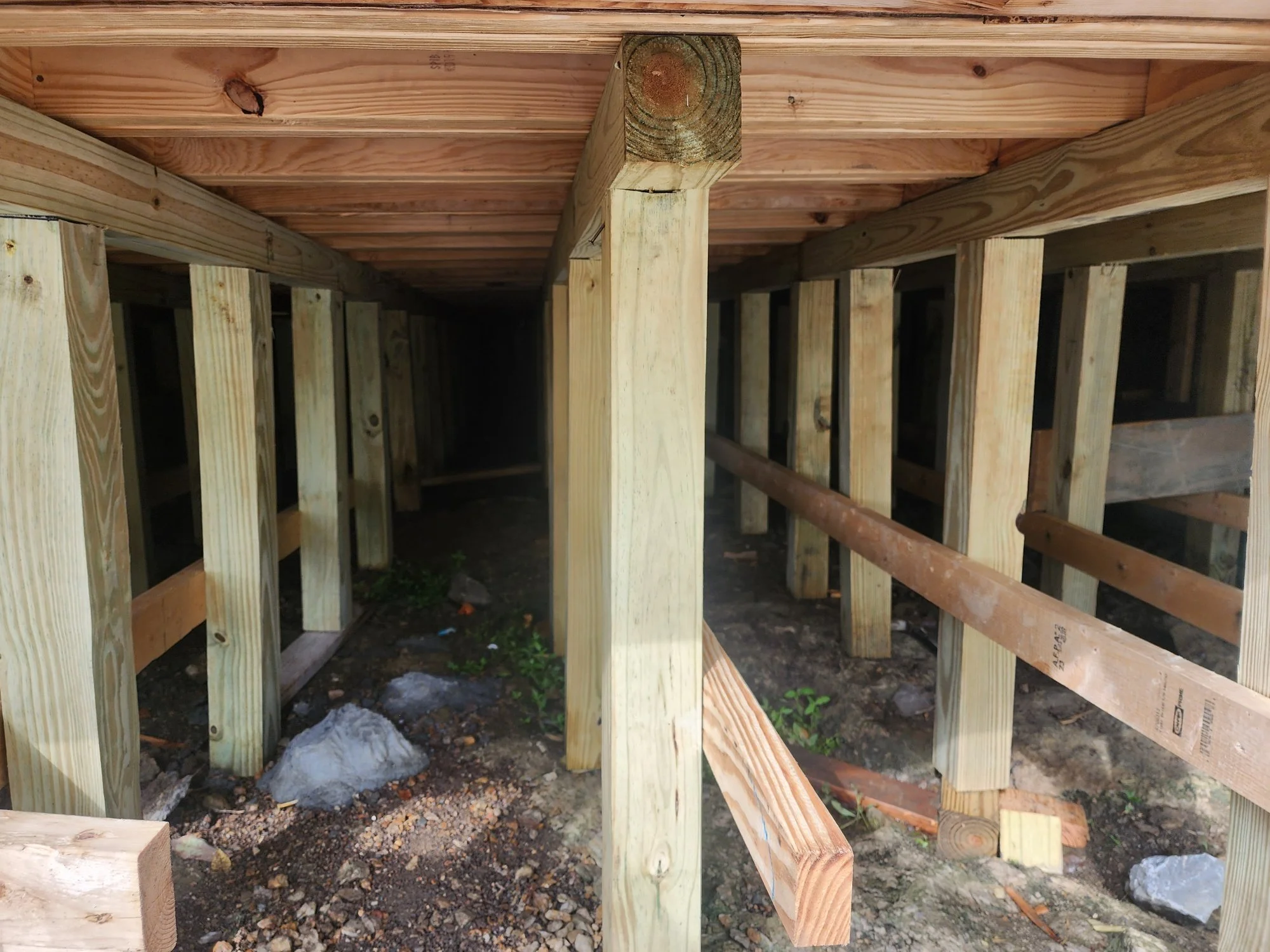 View of a wooden deck foundation with vertical support beams and horizontal joists, showing construction materials and dirt ground underneath.