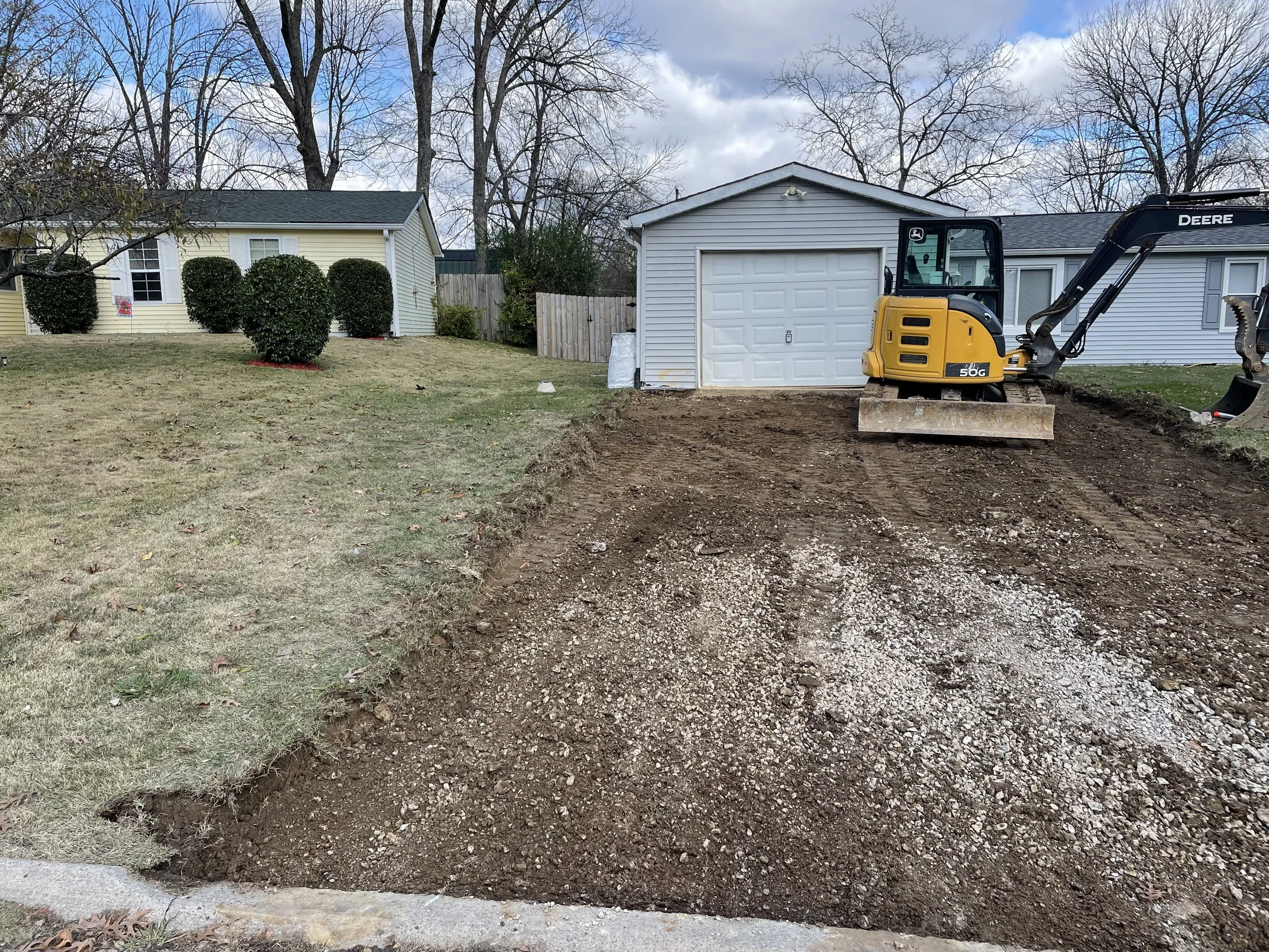 A backyard with a freshly excavated area and a small yellow and black mini excavator on the soil. There are houses with bushes and trees in the background, and a partly cloudy sky overhead.