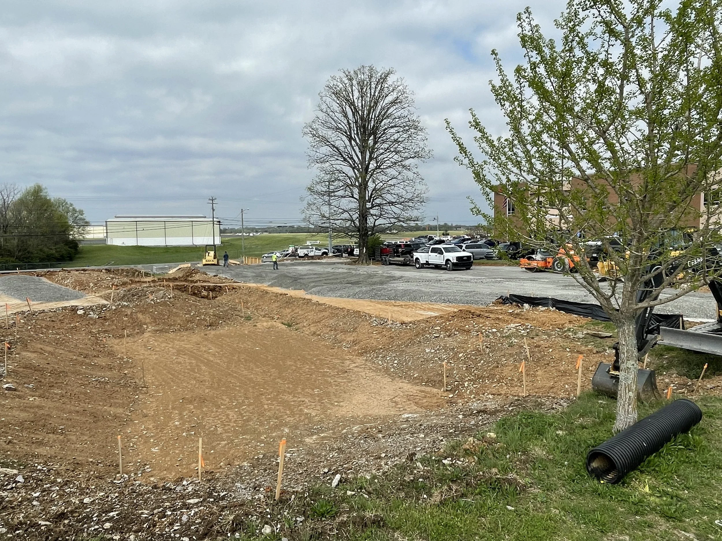 A construction site with dirt and gravel, trees, parked cars, and a building in the background under a cloudy sky.