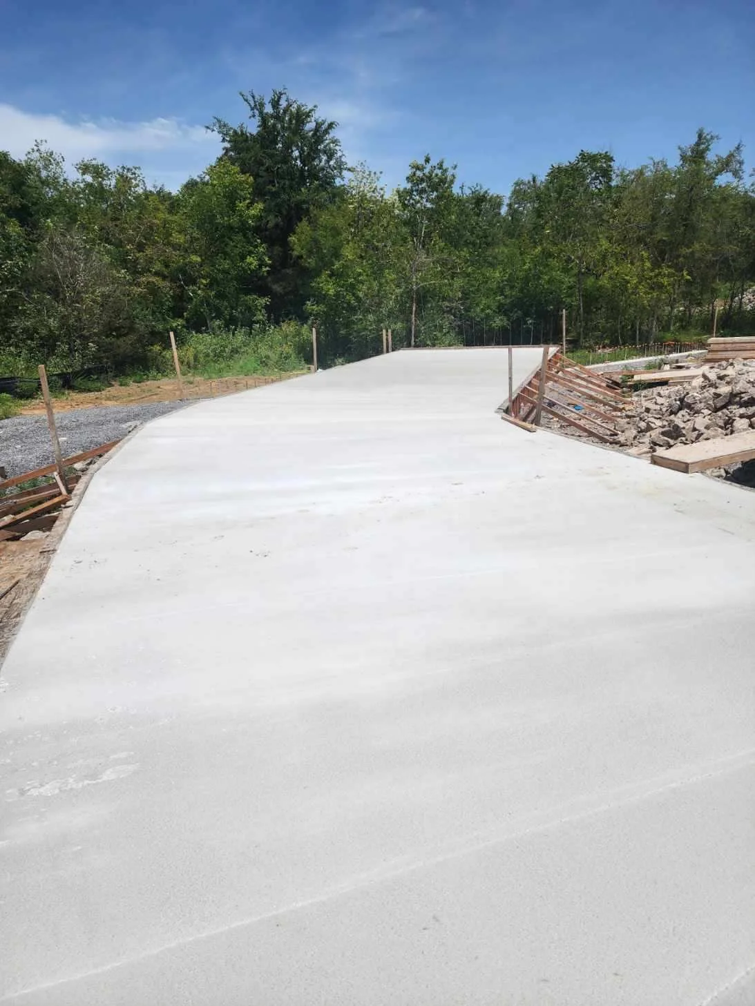 Construction site with newly poured concrete sidewalk and surrounding wooden forms, with trees and a blue sky in the background.