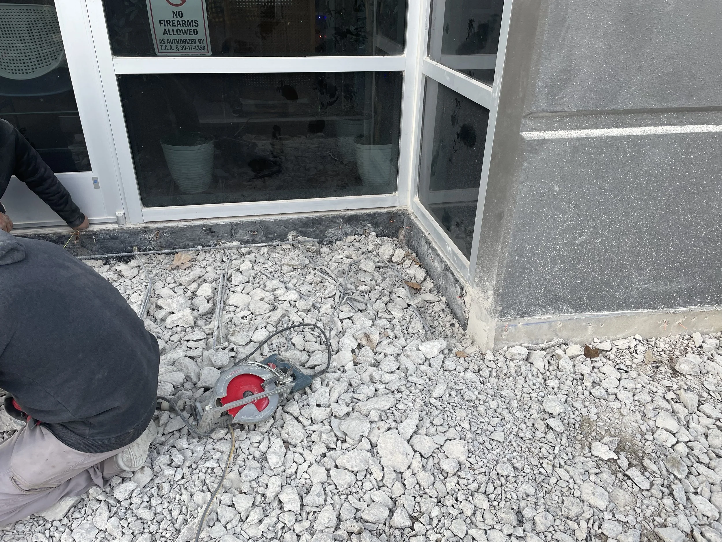 Construction worker kneeling on gravel outside a building with large glass windows, using a circular saw.
