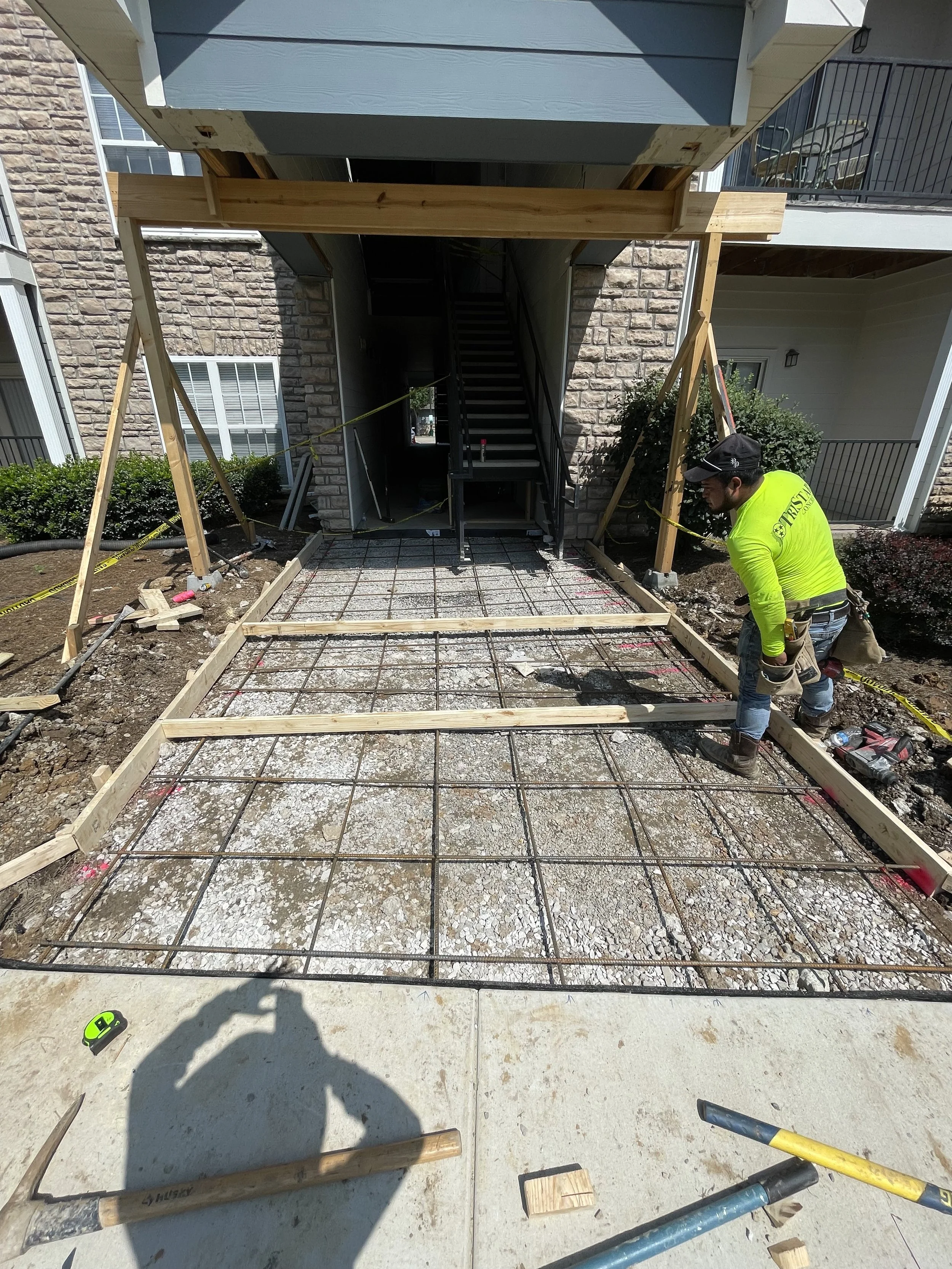 Construction worker wearing a neon yellow shirt pouring concrete over a rebar grid for a new concrete porch at the entrance of an apartment building.