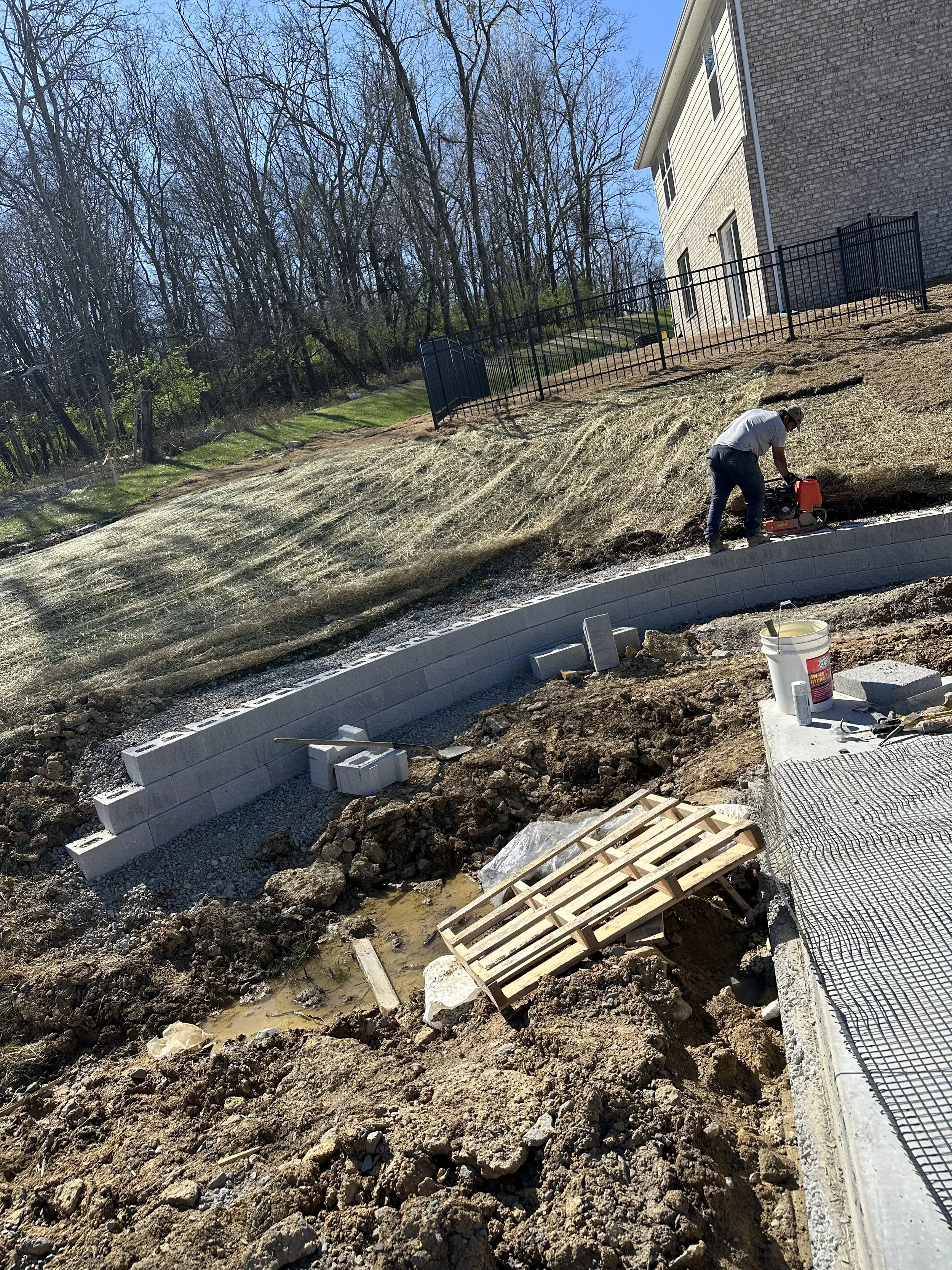 A person working on a construction project laying bricks on a curved retaining wall, with building tools, scattered bricks, and a pallet nearby, on a sloped backyard with a house and trees in the background.