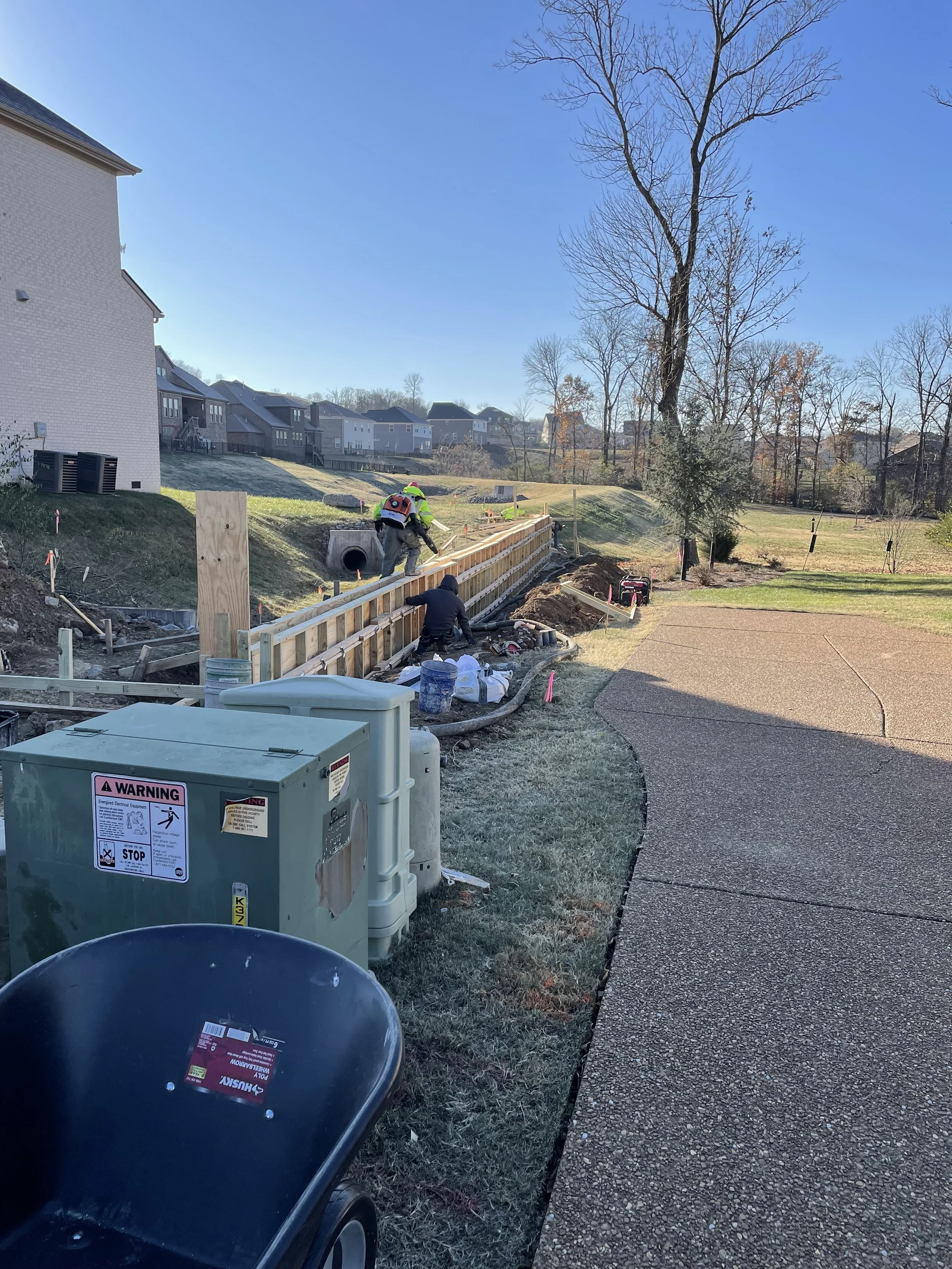 Construction workers building a retaining wall along a curved walking path in a residential area on a clear, sunny day.