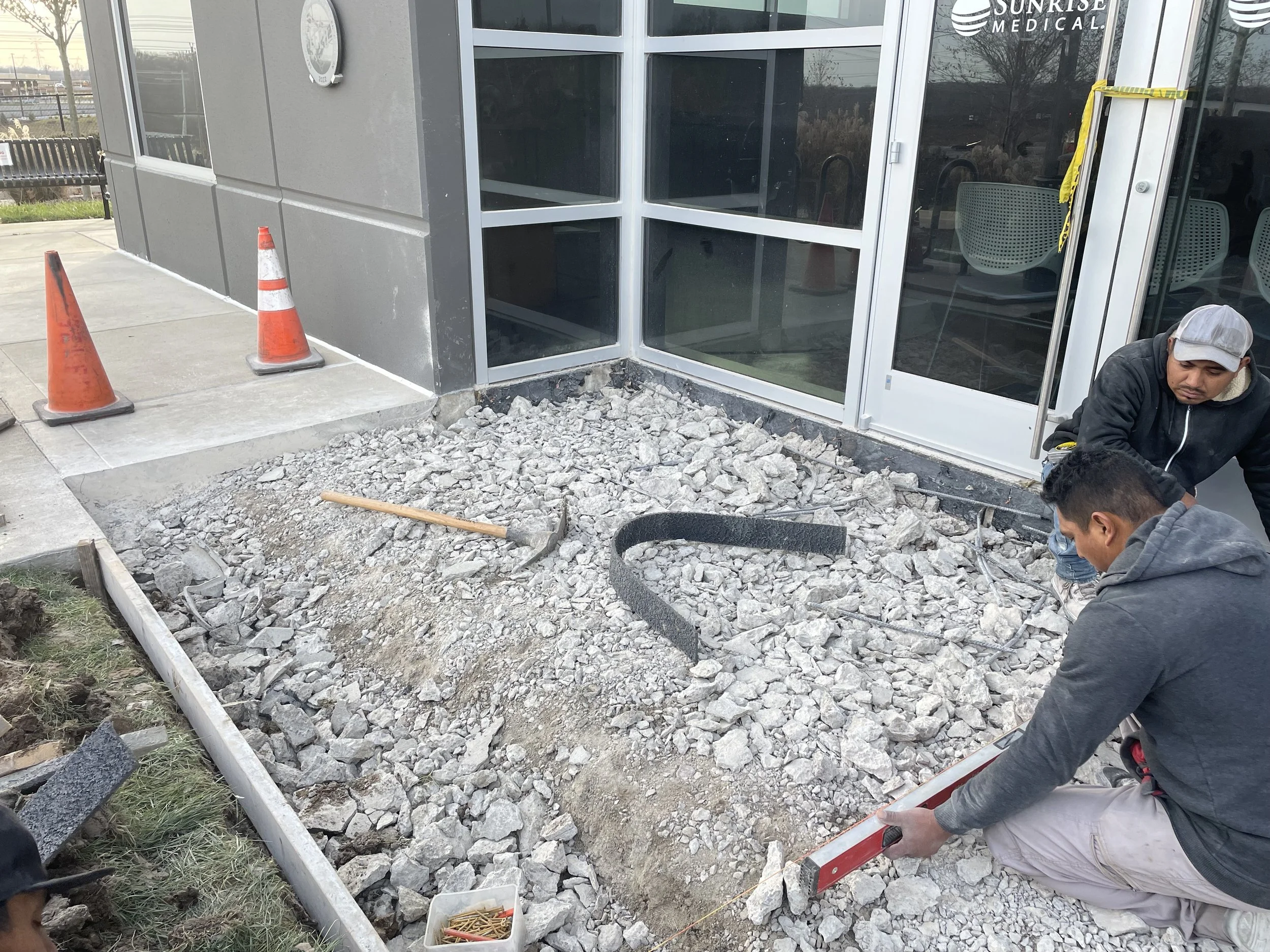 Construction workers leveling gravel in front of a building with large glass windows and orange safety cones nearby.
