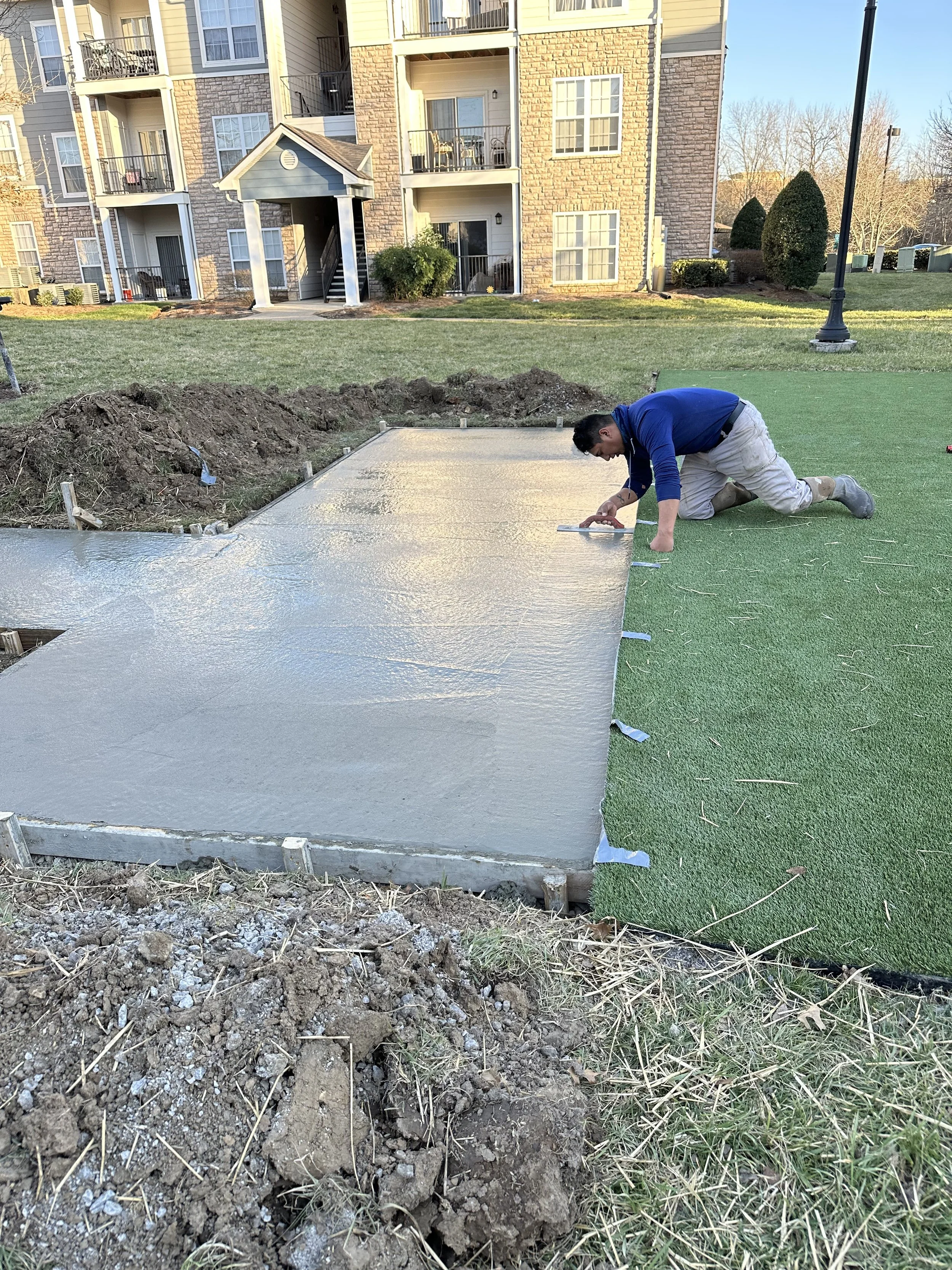 A man kneeling on the ground, smoothing a freshly poured concrete slab outside an apartment building, with green artificial grass adjacent to the concrete.