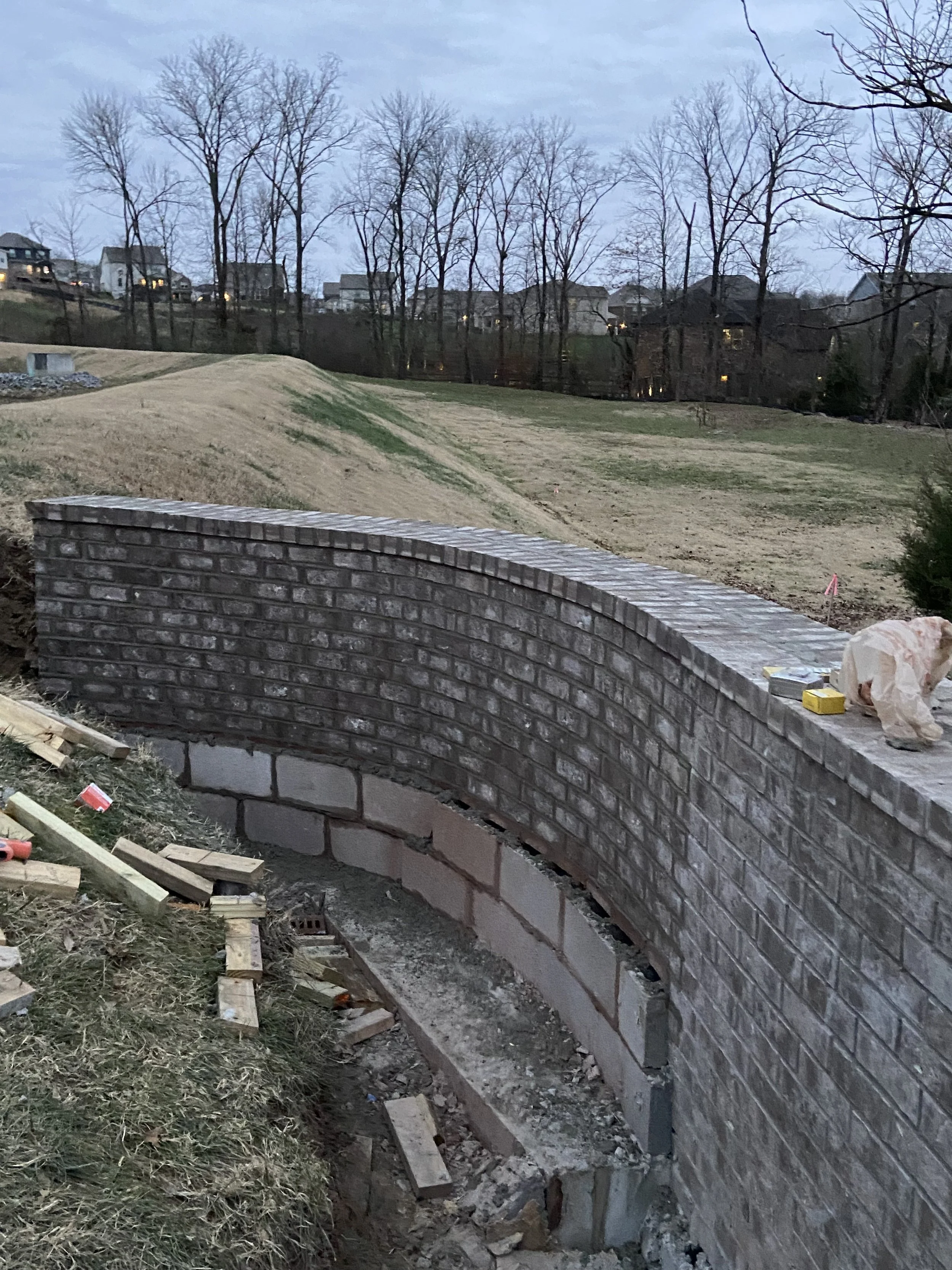 Construction of a curved brick wall in a backyard with grassy hills and leafless trees in the background.