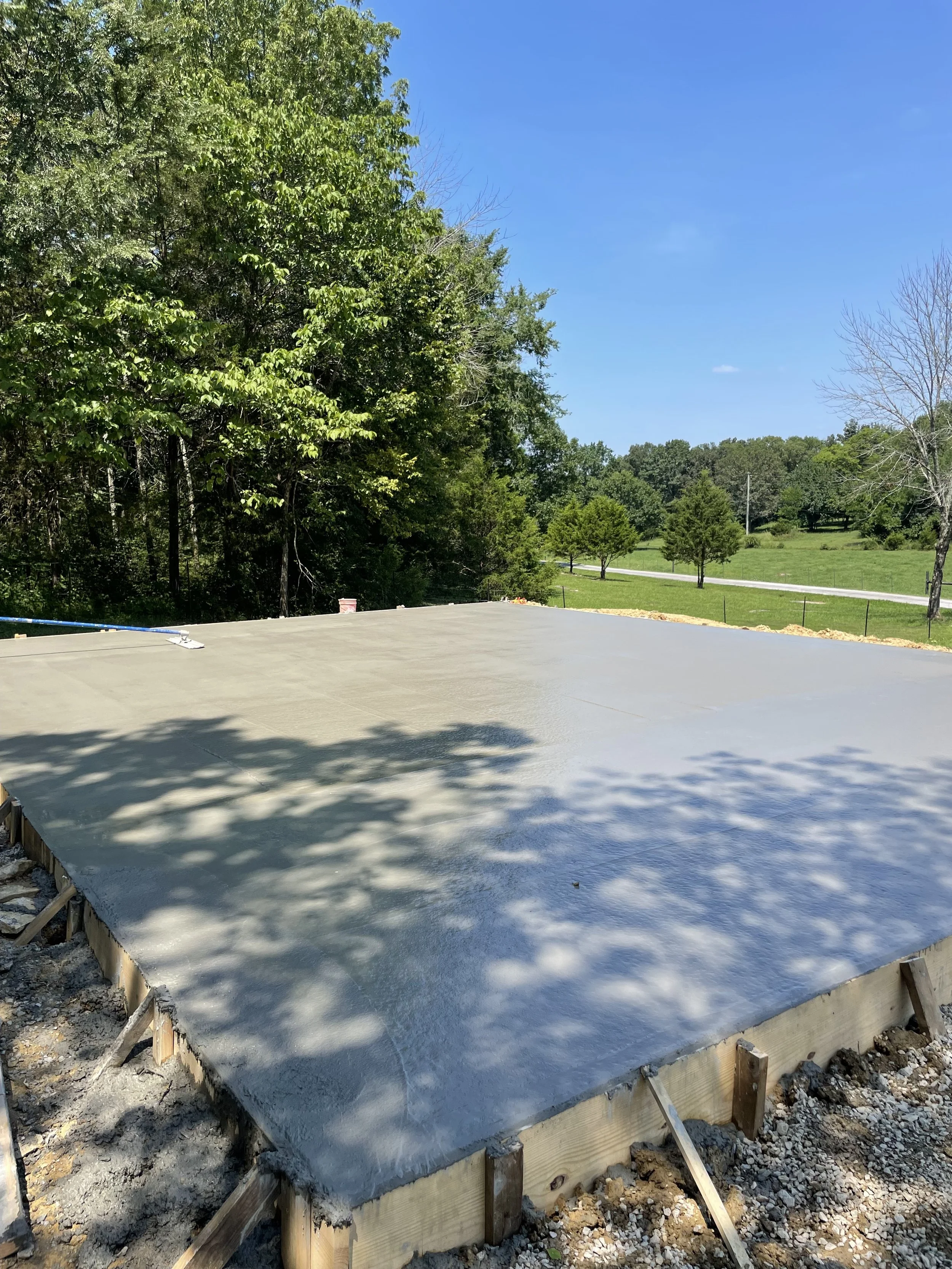 Freshly poured concrete slab foundation on a construction site, surrounded by trees and grass under a clear blue sky.