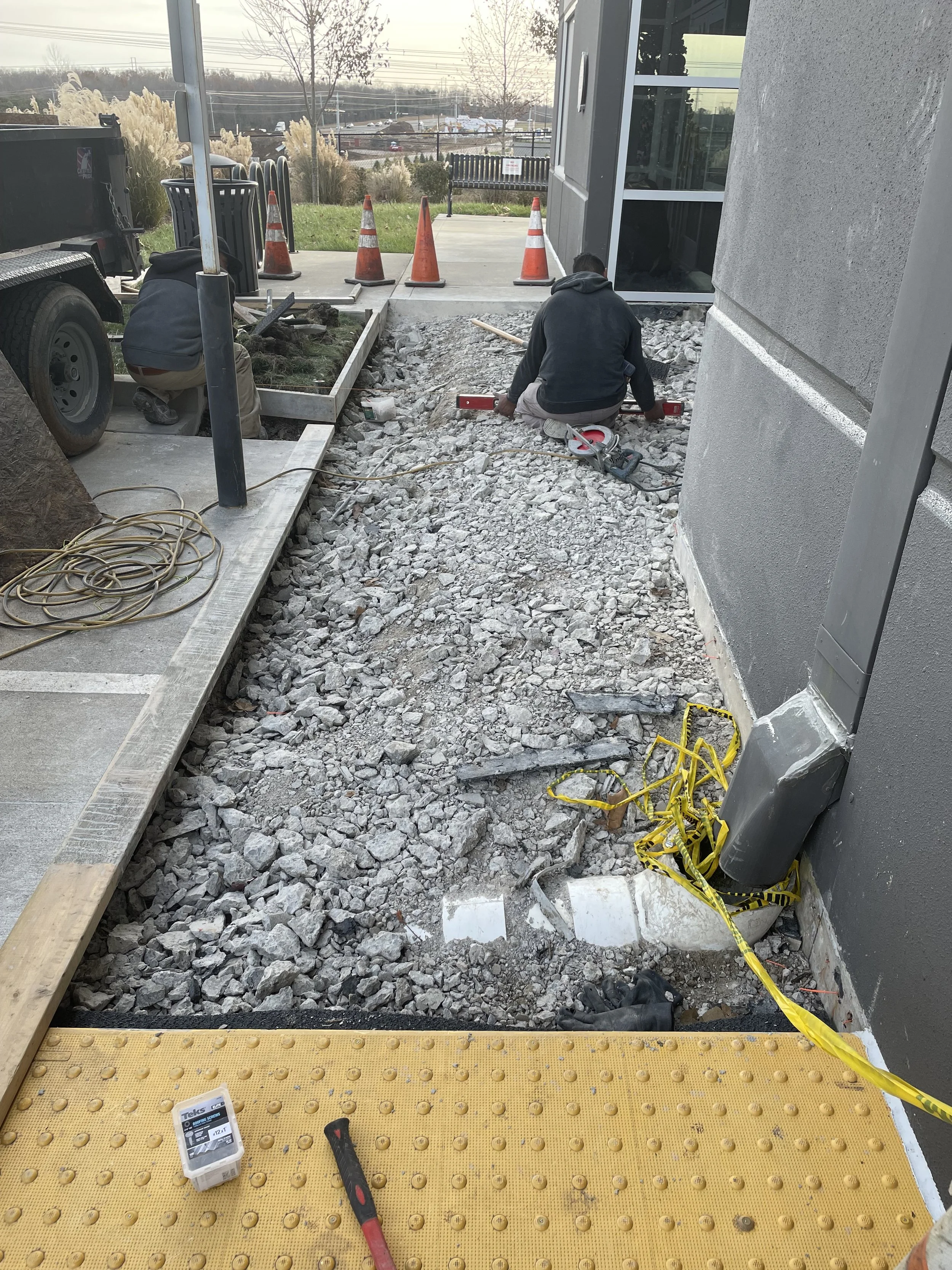 Construction workers repairing a sidewalk outside a building, with orange traffic cones, tools, and yellow caution tape present.