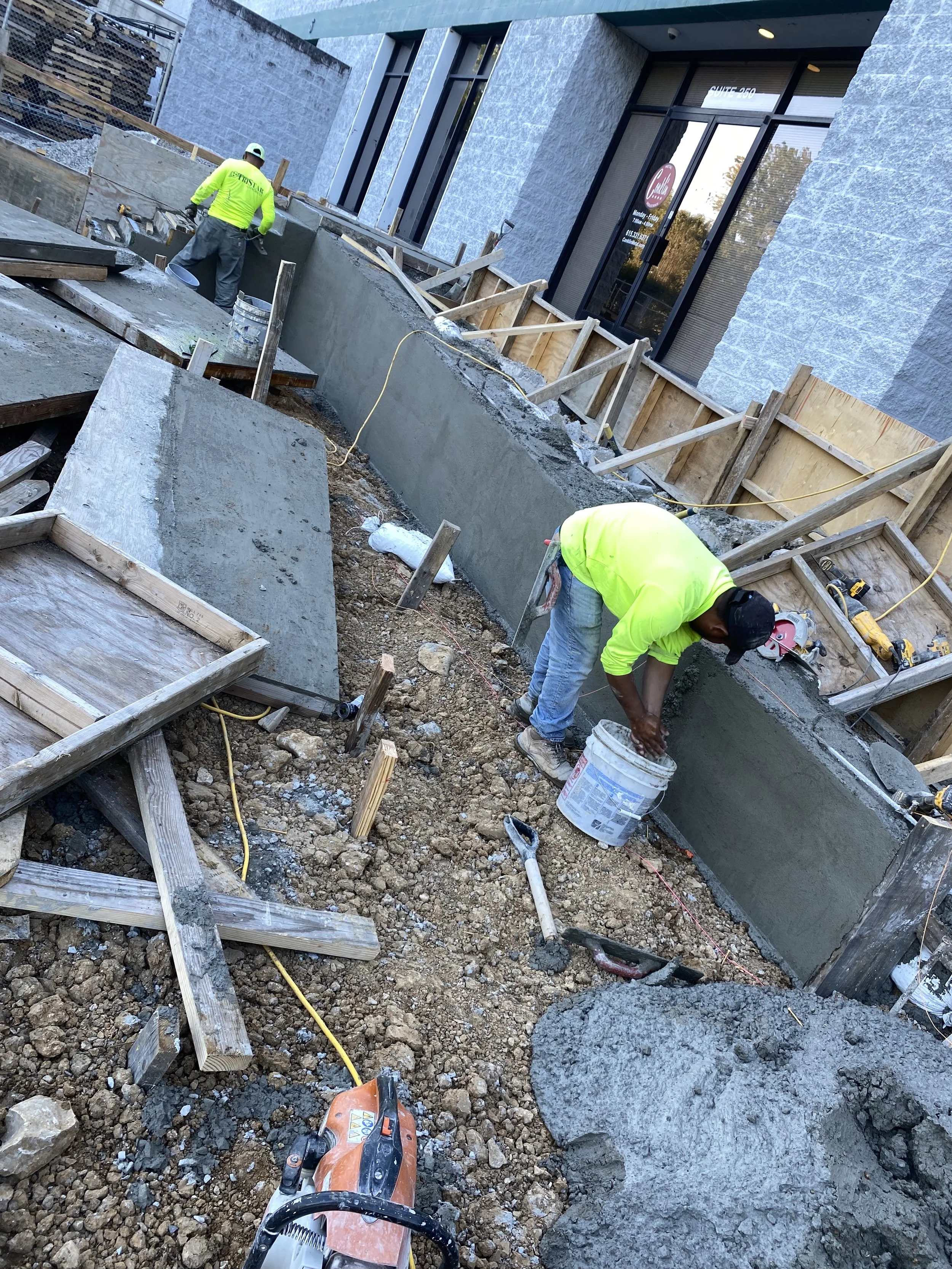 Workers in bright yellow safety shirts constructing a concrete building foundation, with one worker leveling concrete and another preparing surface with tools on a dirt ground.