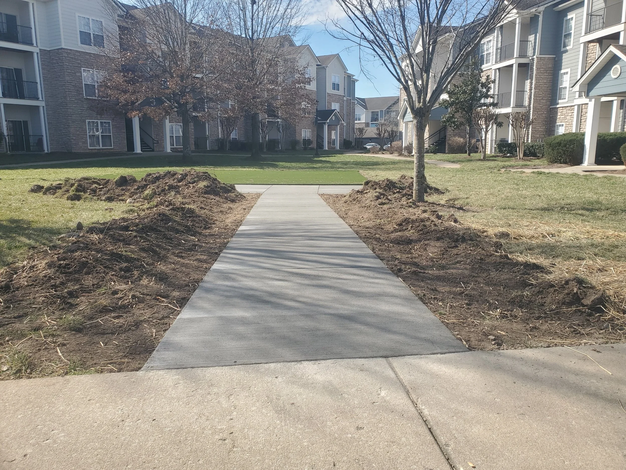 Newly paved sidewalk in apartment complex courtyard, bordered by dirt and grass, with trees and residential buildings in the background.