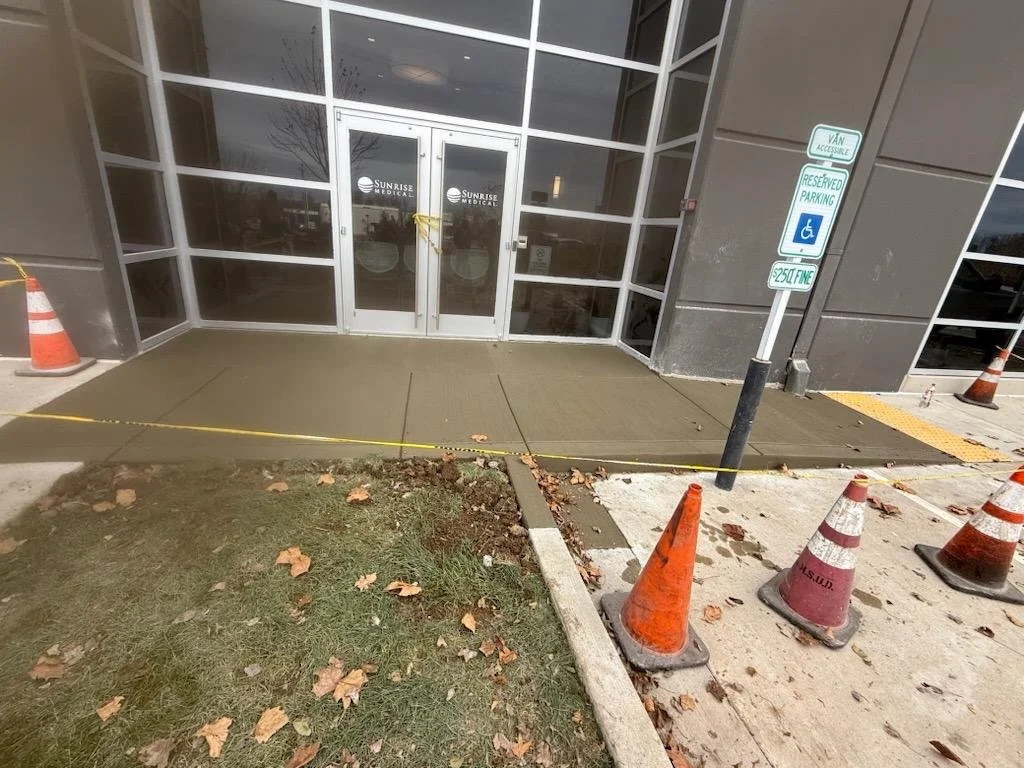 Newly installed wheelchair accessible ramp in front of Sunrise Medical office, surrounded by orange traffic cones and caution tape, with reserved parking signs.