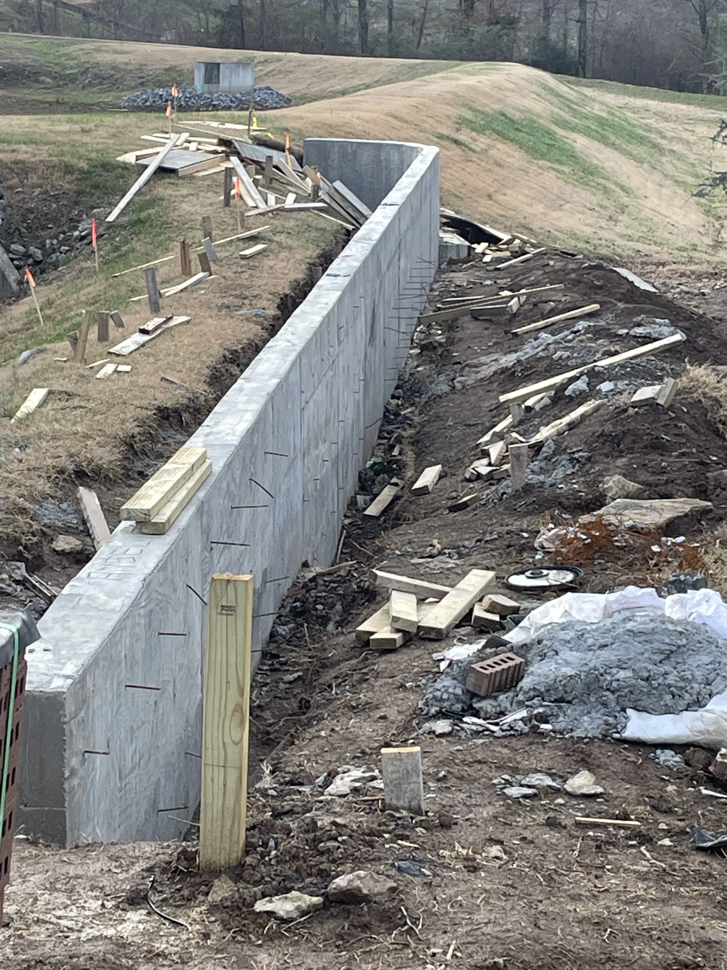 Construction site of a concrete wall, with scattered wooden planks, bricks, and construction tools on uneven dirt ground, with a hillside and trees in the background.