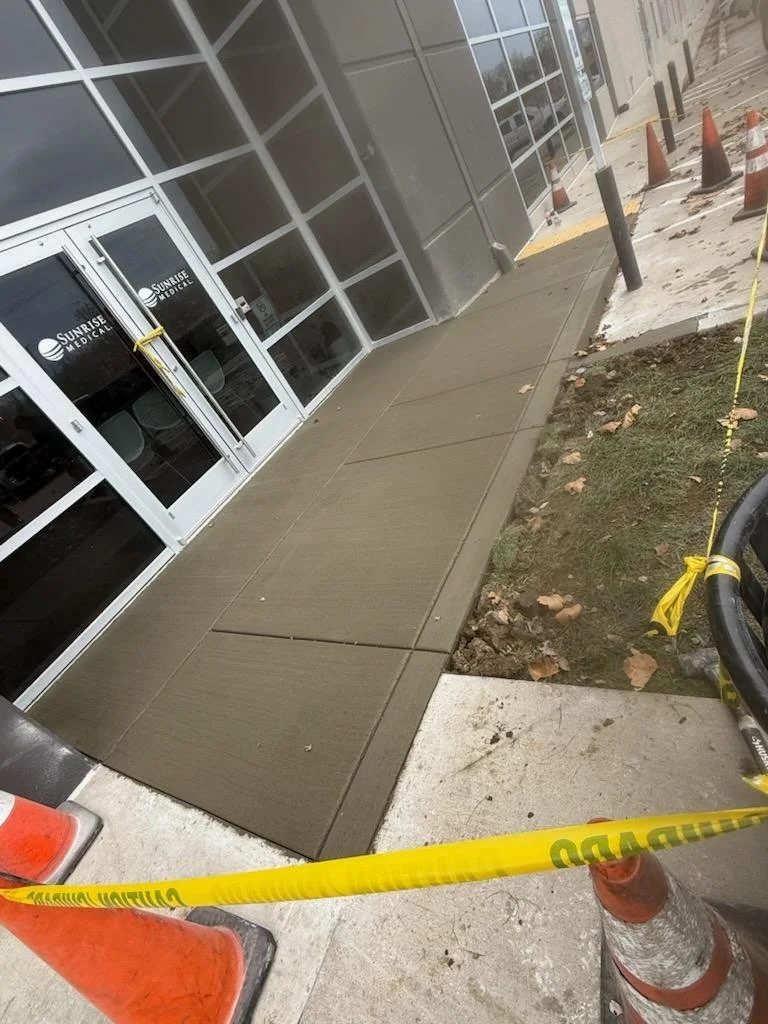 Newly installed concrete sidewalk outside a medical building with safety cones and caution tape around the work area