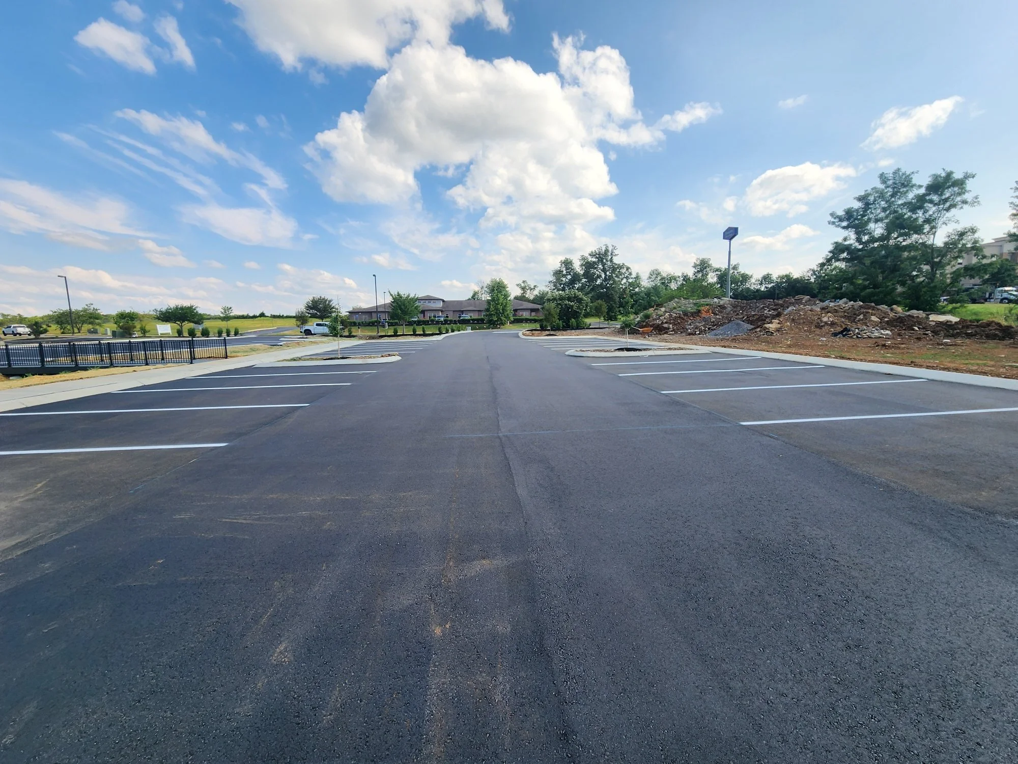 Empty parking lot with freshly paved asphalt and white parking space lines under a partly cloudy sky with green trees in the background.