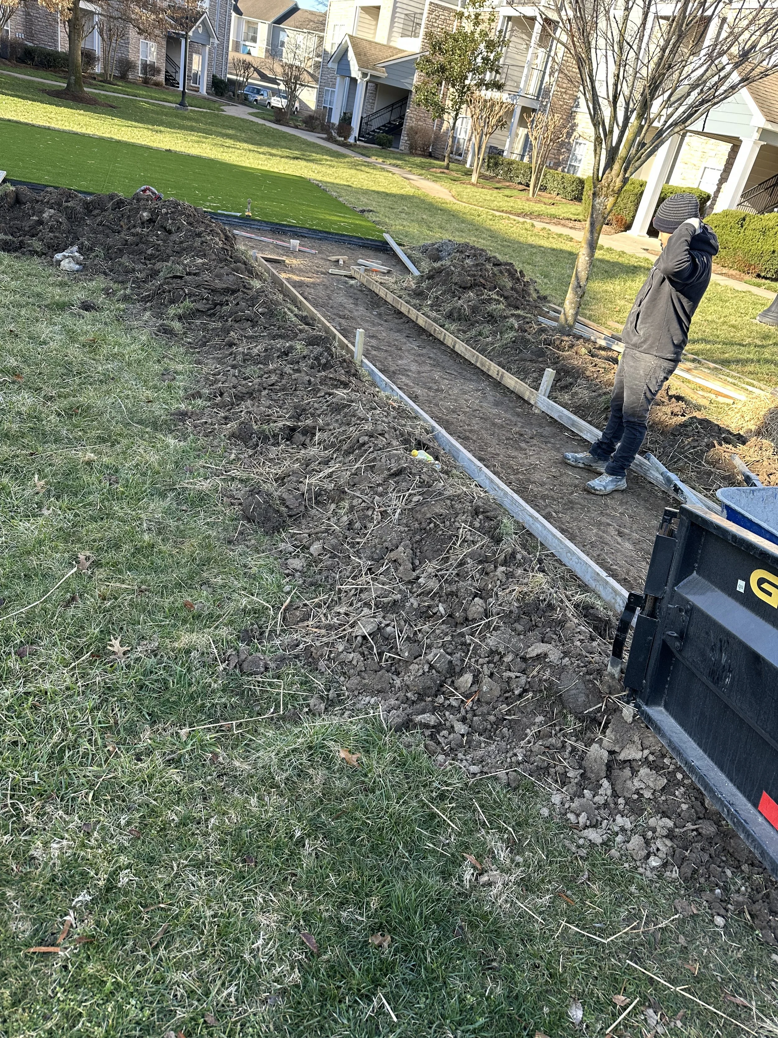 A person standing next to a freshly dug and framed sidewalk or garden bed in a residential area with grass, trees, and houses in the background.