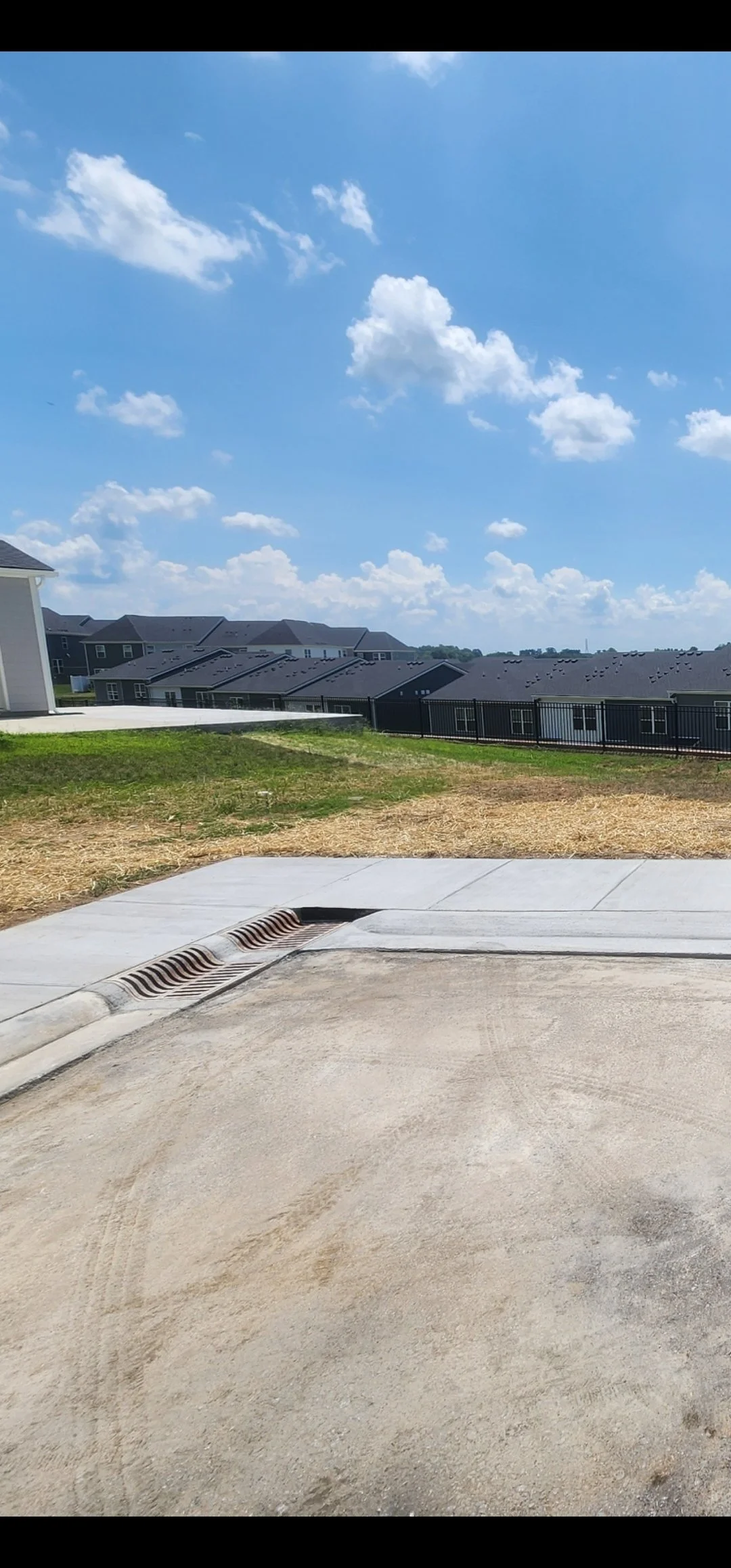 Empty street view with a concrete curb, a drainage grate, a grassy area, apartment buildings in the distance, and a blue sky with fluffy clouds.