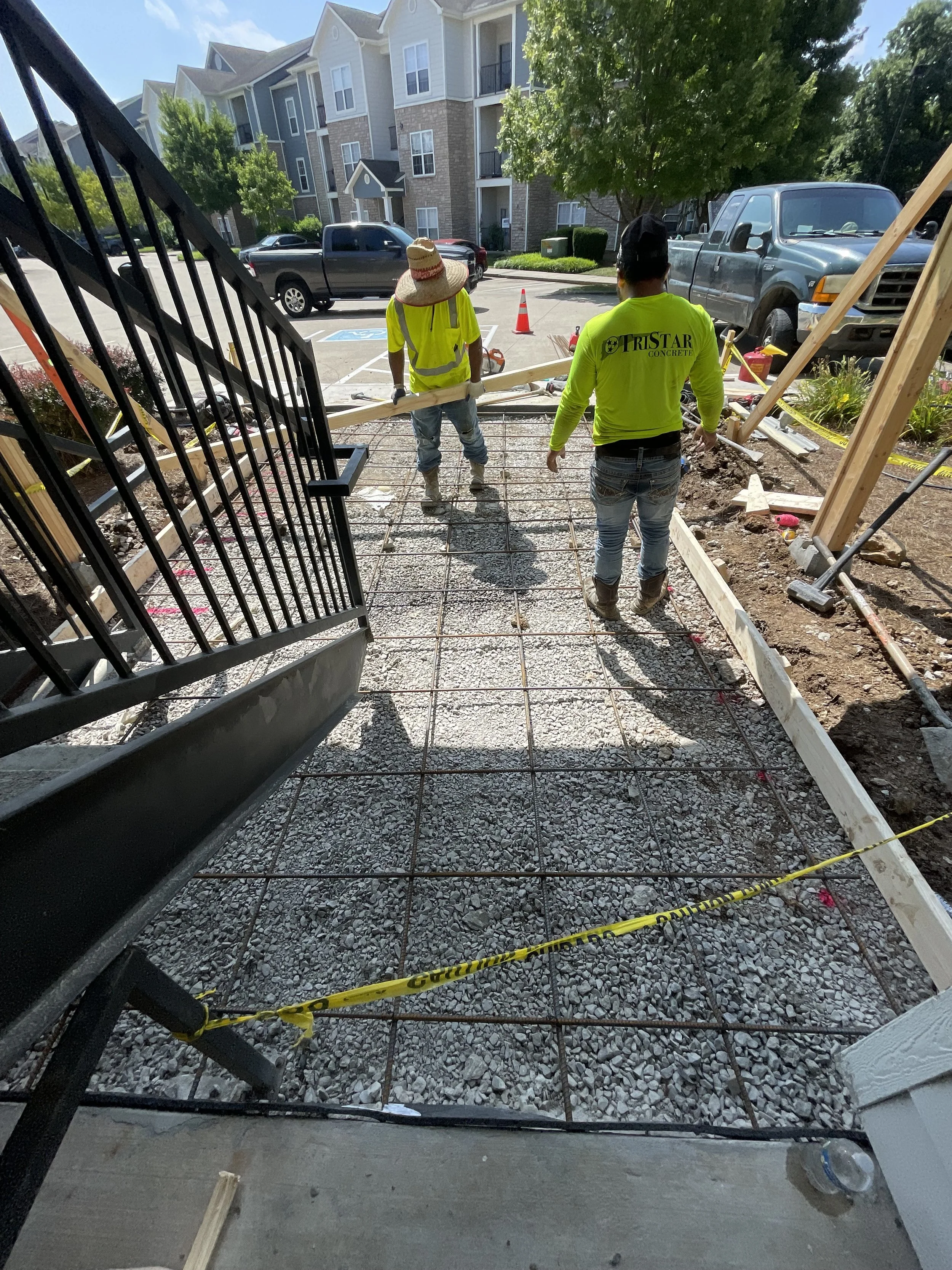 Two construction workers are working on pouring a concrete sidewalk with rebar reinforcement in a residential area. One worker is wearing a large straw hat and a high-visibility vest, while the other has on a bright yellow long-sleeve shirt with the