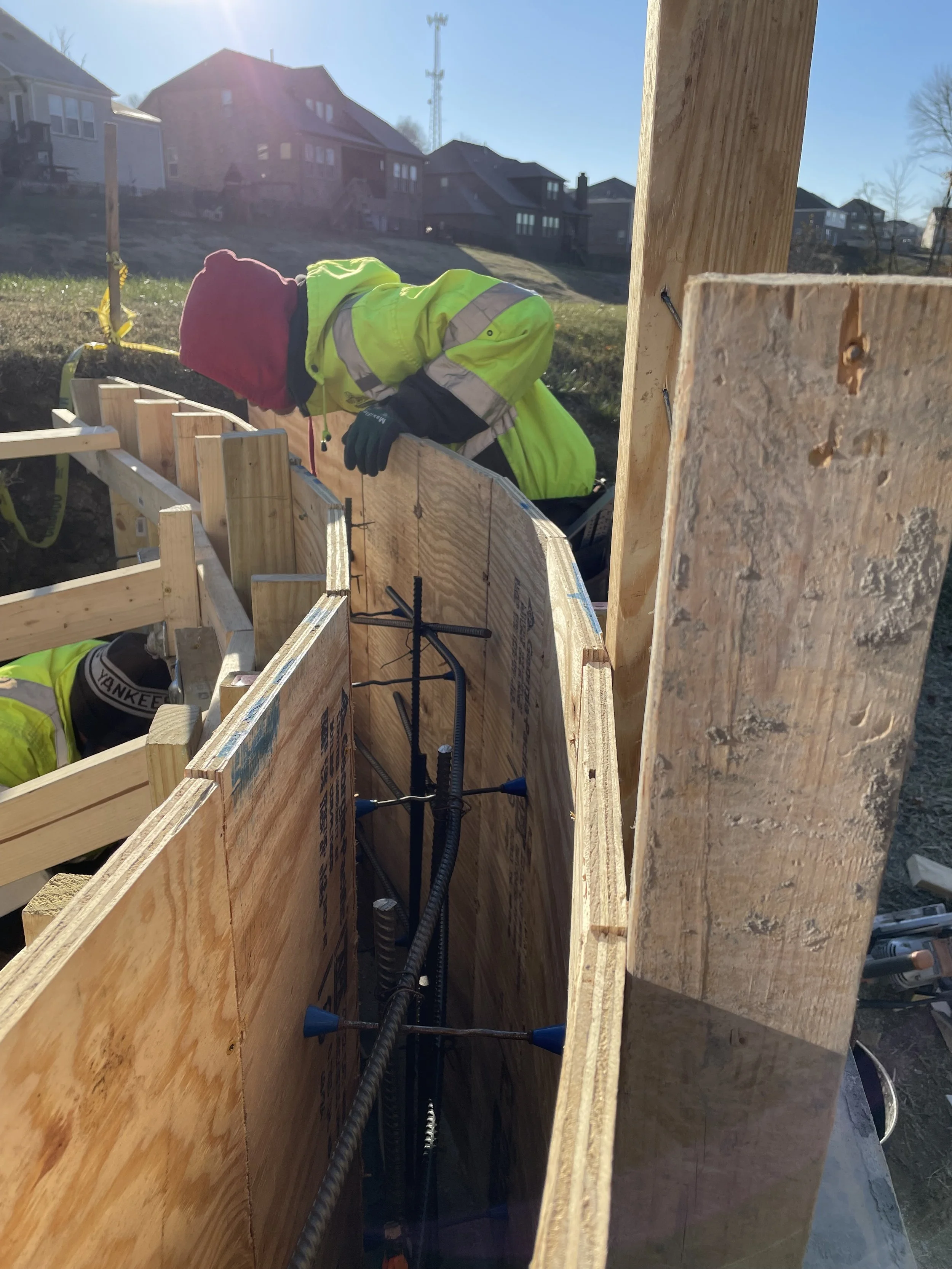 Construction worker in a neon yellow jacket and red beanie working on a building foundation during daylight, with wooden framing and rebar visible.