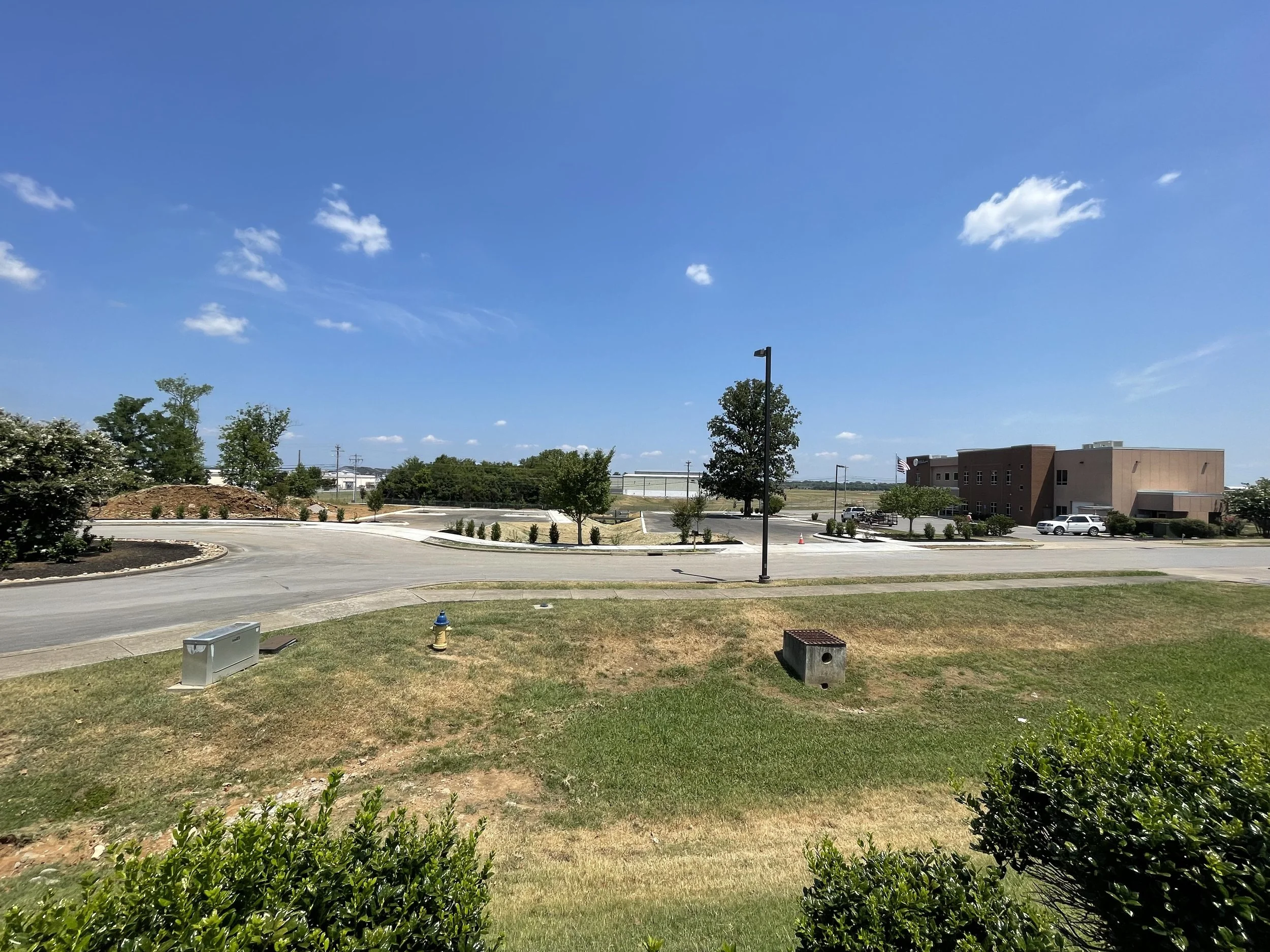 Empty parking lot with a few trees, a building in the background, and a bright blue sky with scattered clouds.